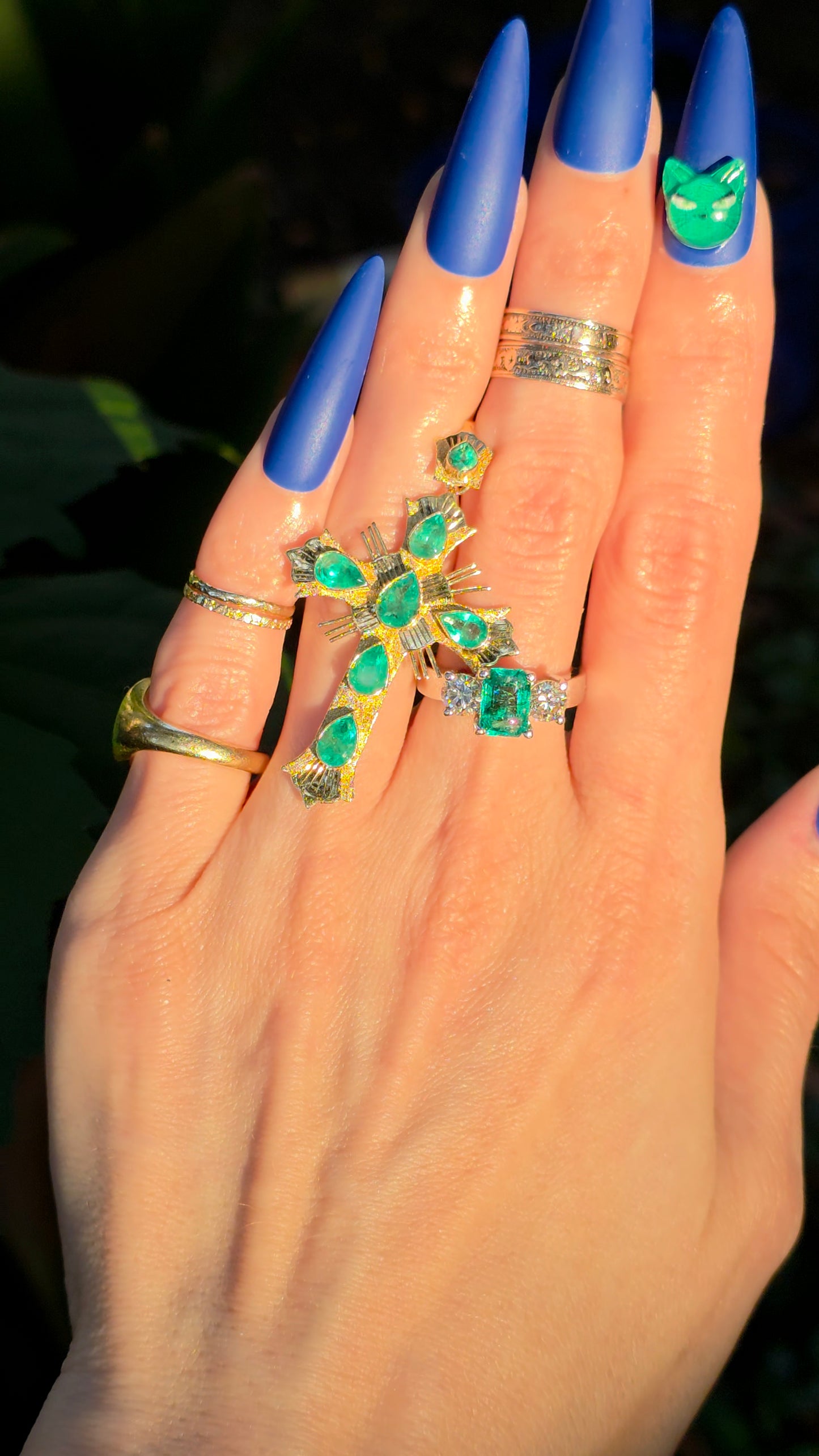 A close-up of a hand with long blue manicured nails wearing multiple rings, including a prominent gold ring with green gemstones and a smaller matching ring on another finger.