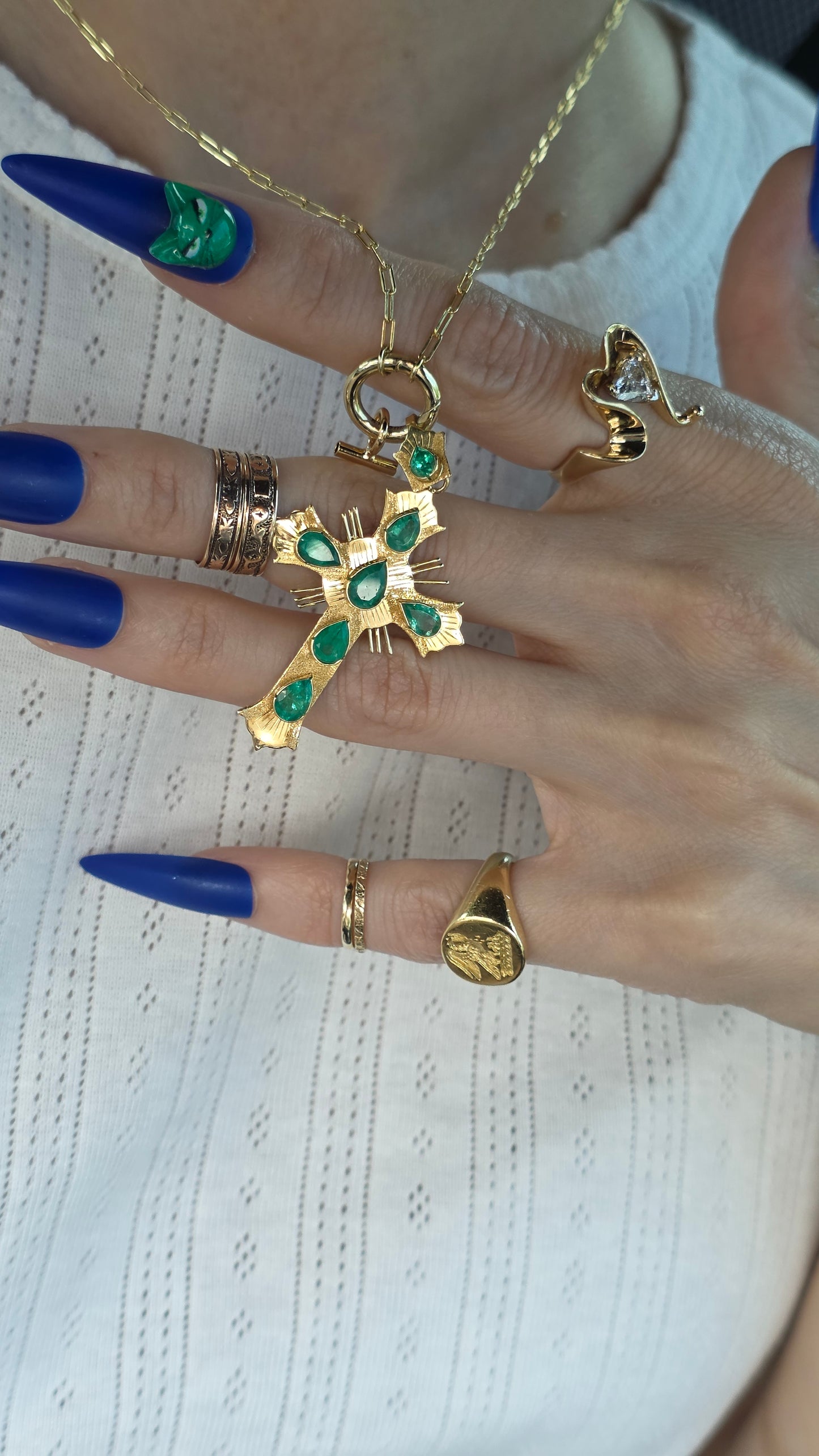 A close-up of a hand wearing multiple gold rings, including a prominent cross ring with green stones, a snake ring, and a signet-style ring, paired with blue stiletto nails against a white textured background.