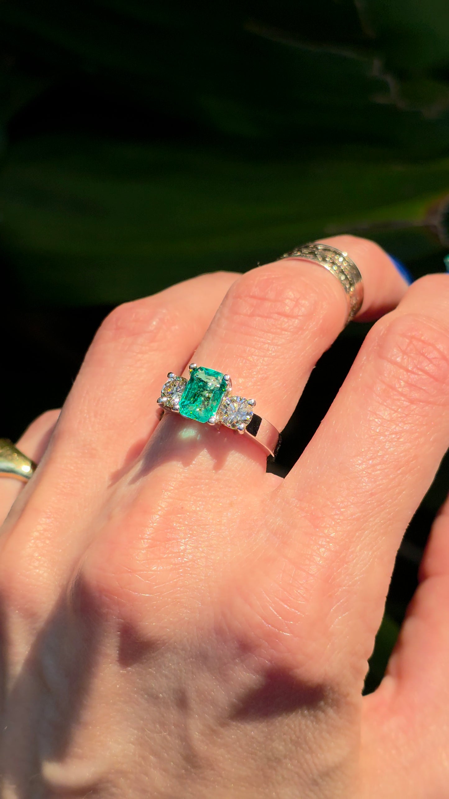 A close-up of a hand wearing a silver ring with a rectangular green emerald-cut center stone and small side stones, photographed outdoors with a dark green leafy background.