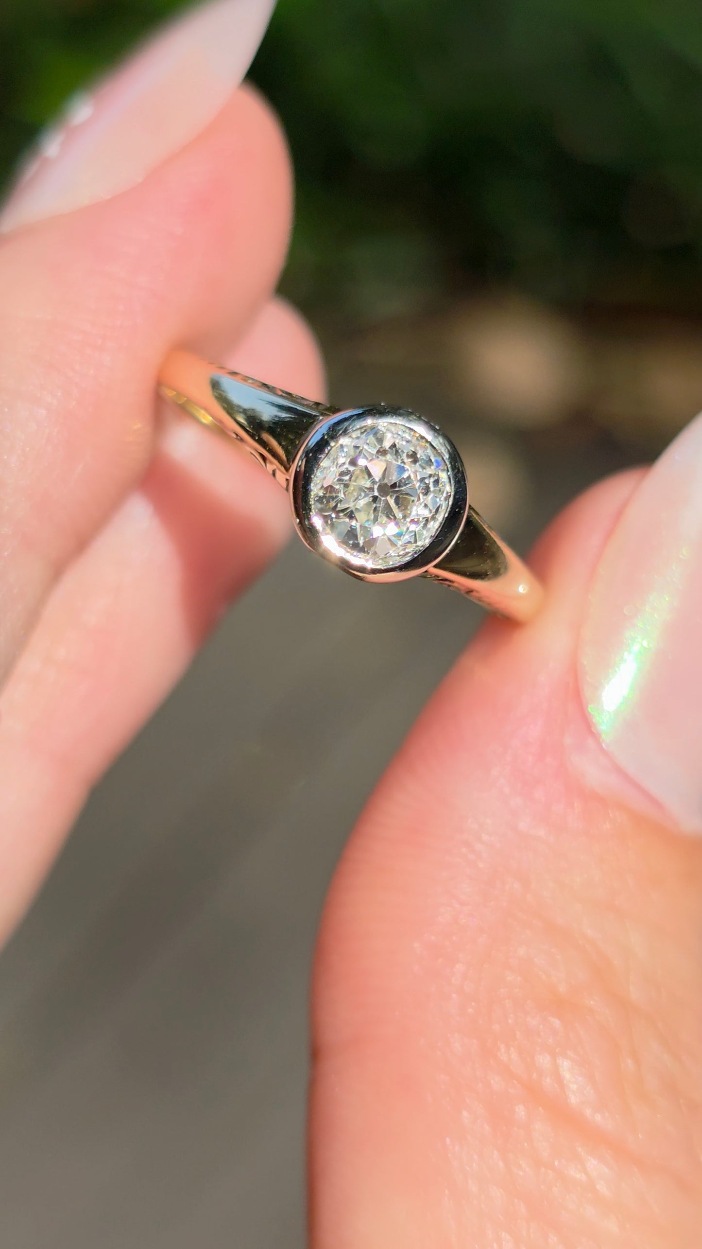 A close-up of a hand holding a gold ring with a round-cut clear gemstone. The ring features a yellow gold band and a bezel-set round diamond, photographed against a blurred outdoor background.