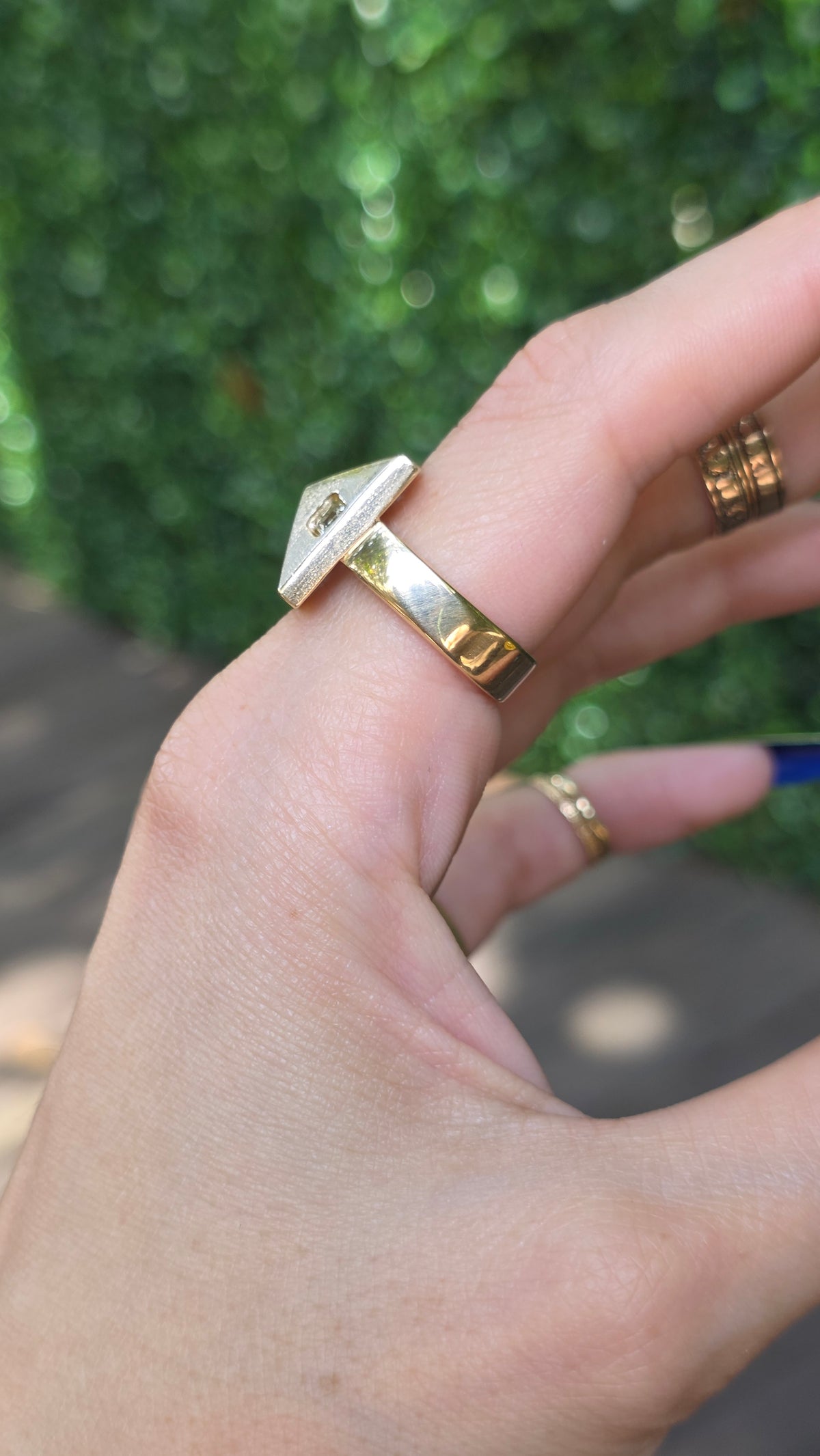 A close-up of a hand wearing several rings, including a prominent gold band with a white rectangular inlay and smaller gold rings, against a blurred green outdoor background.