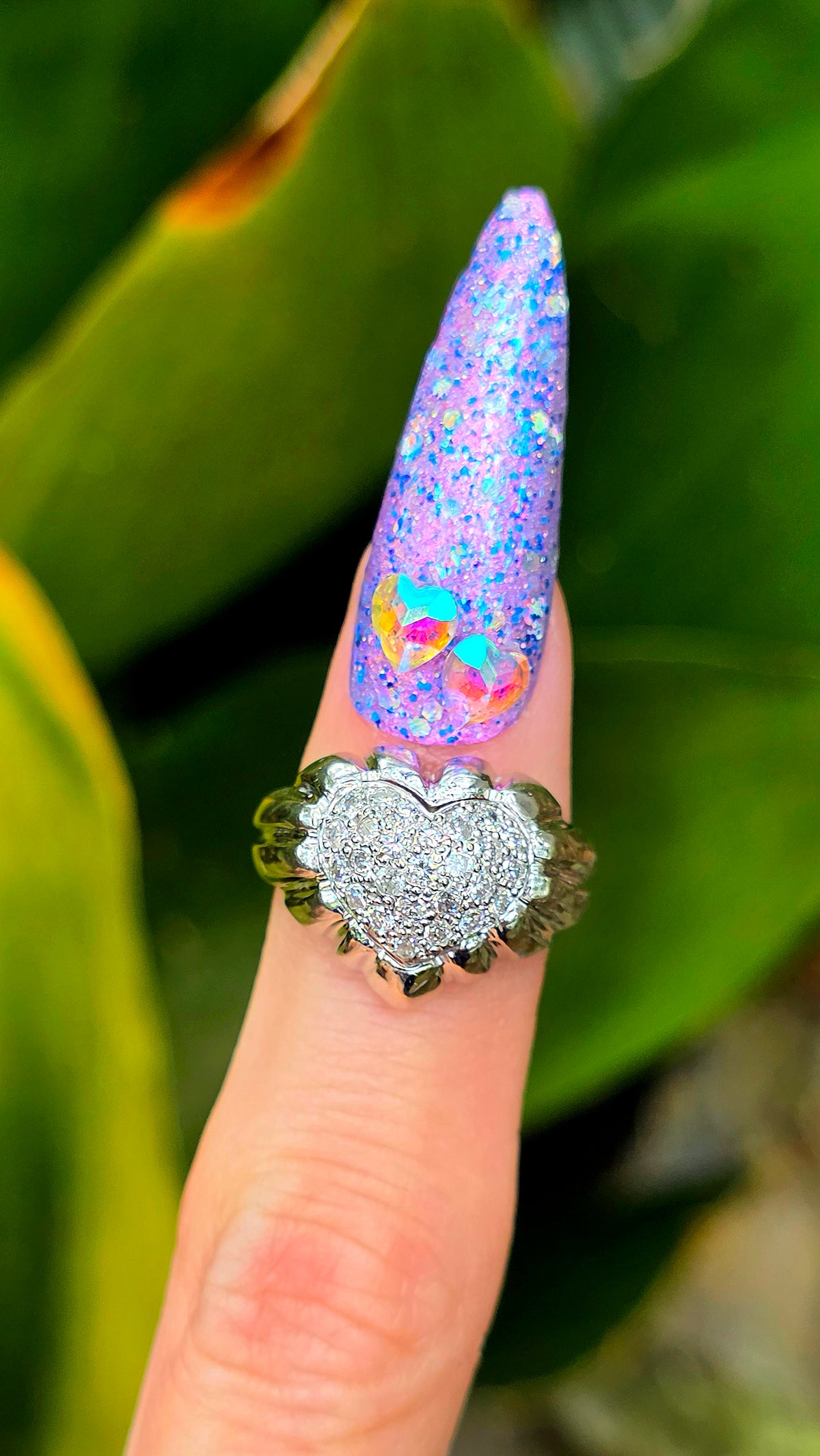 Decorative ring with a heart design on a finger against a green leafy background