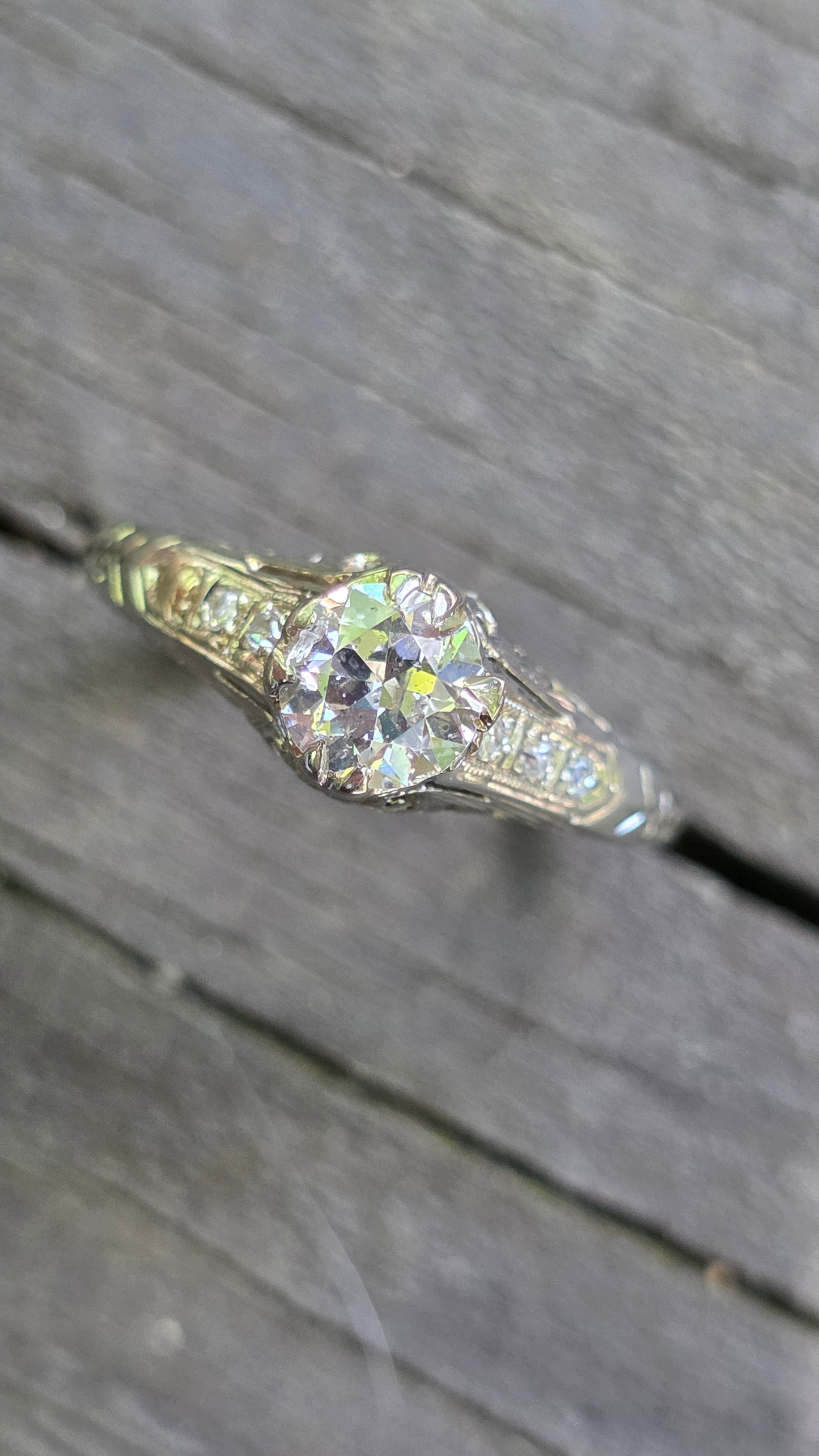 An antique-style diamond ring with a round center stone and smaller side stones on a weathered gray wooden surface.