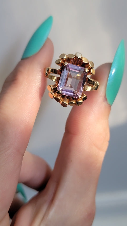 Gold ring with a purple gemstone held between fingers against a neutral background