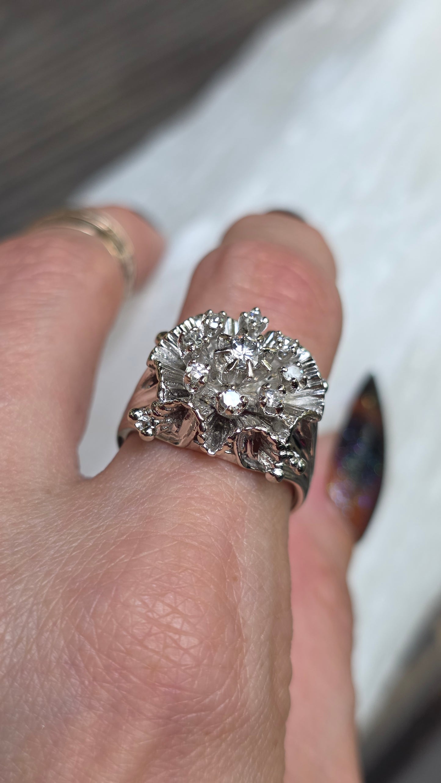 A close-up of a hand wearing a large silver-toned ring with a prominent round cluster of clear gemstones, photographed against a light neutral background.