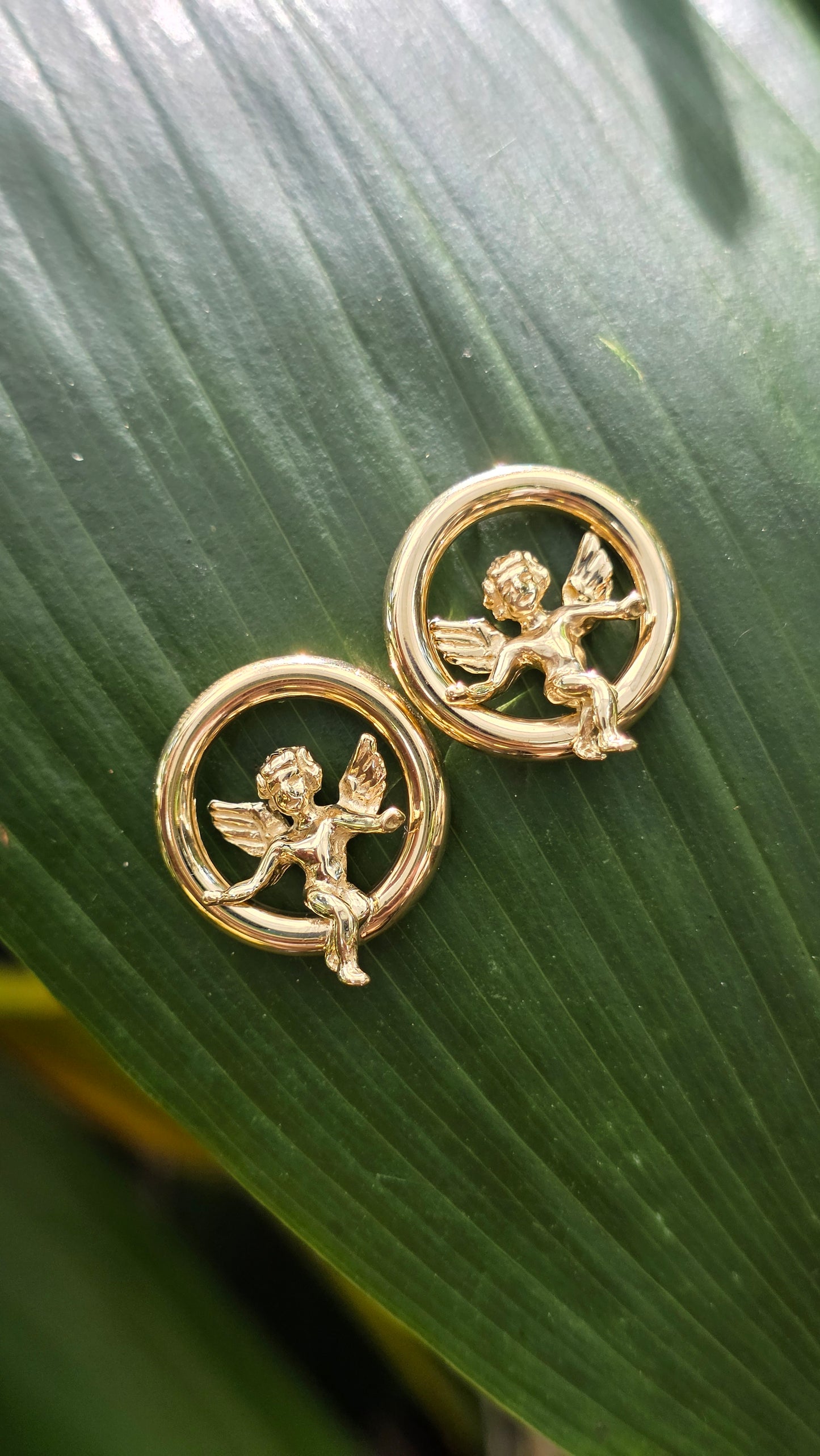 A pair of round vintage 14k yellow gold earrings featuring detailed cherub angels in mid-flight inside polished gold circular frames, resting on a green leaf background.