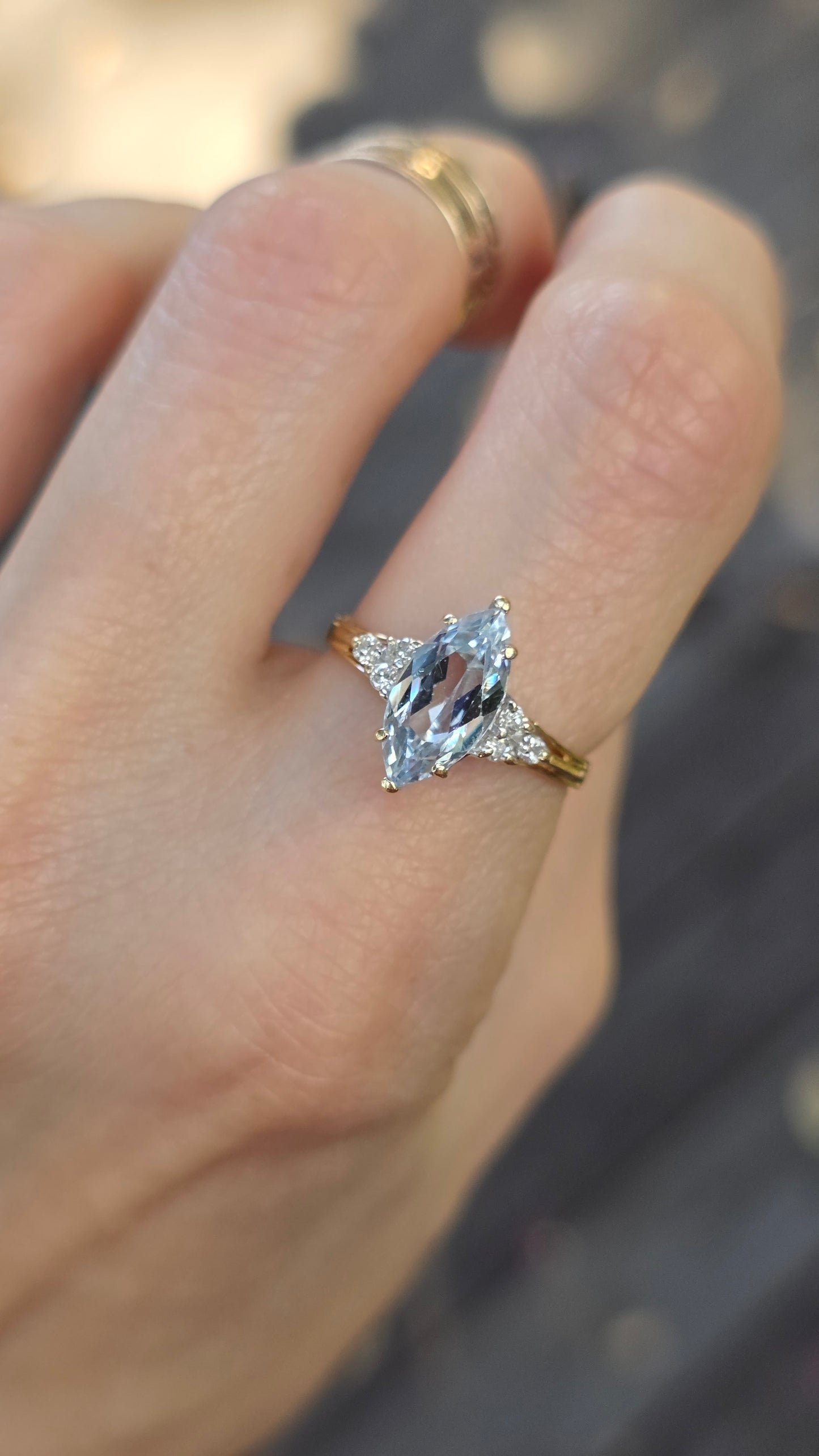 A close-up of a hand wearing a gold ring with a marquise-cut clear gemstone and smaller side stones, with a plain gold band visible on the adjacent finger.
