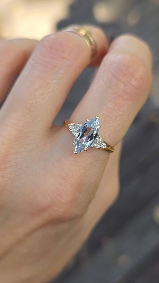 A close-up of a hand wearing a gold ring with a marquise-cut clear gemstone and smaller side stones, with a plain gold band visible on the adjacent finger.
