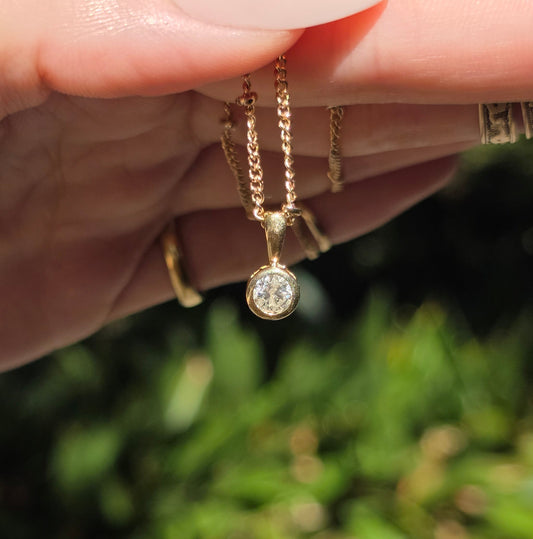 A close-up of a hand holding a delicate gold chain necklace with a small round clear gemstone pendant, set against a blurred green outdoor background.