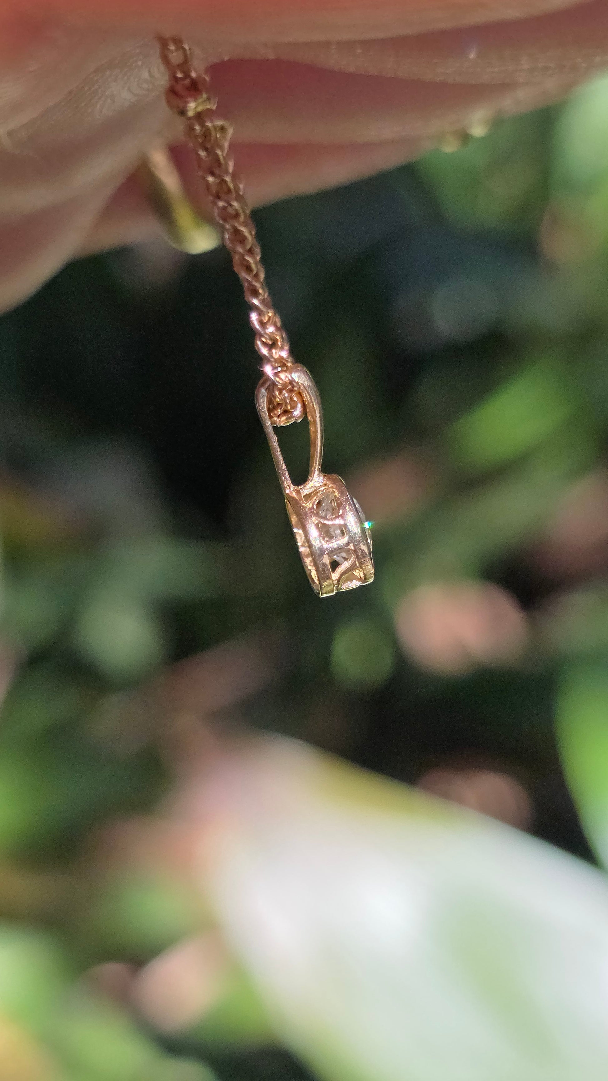 A close-up of a delicate rose gold chain necklace with a small padlock pendant, held by a hand against a blurred green outdoor background.