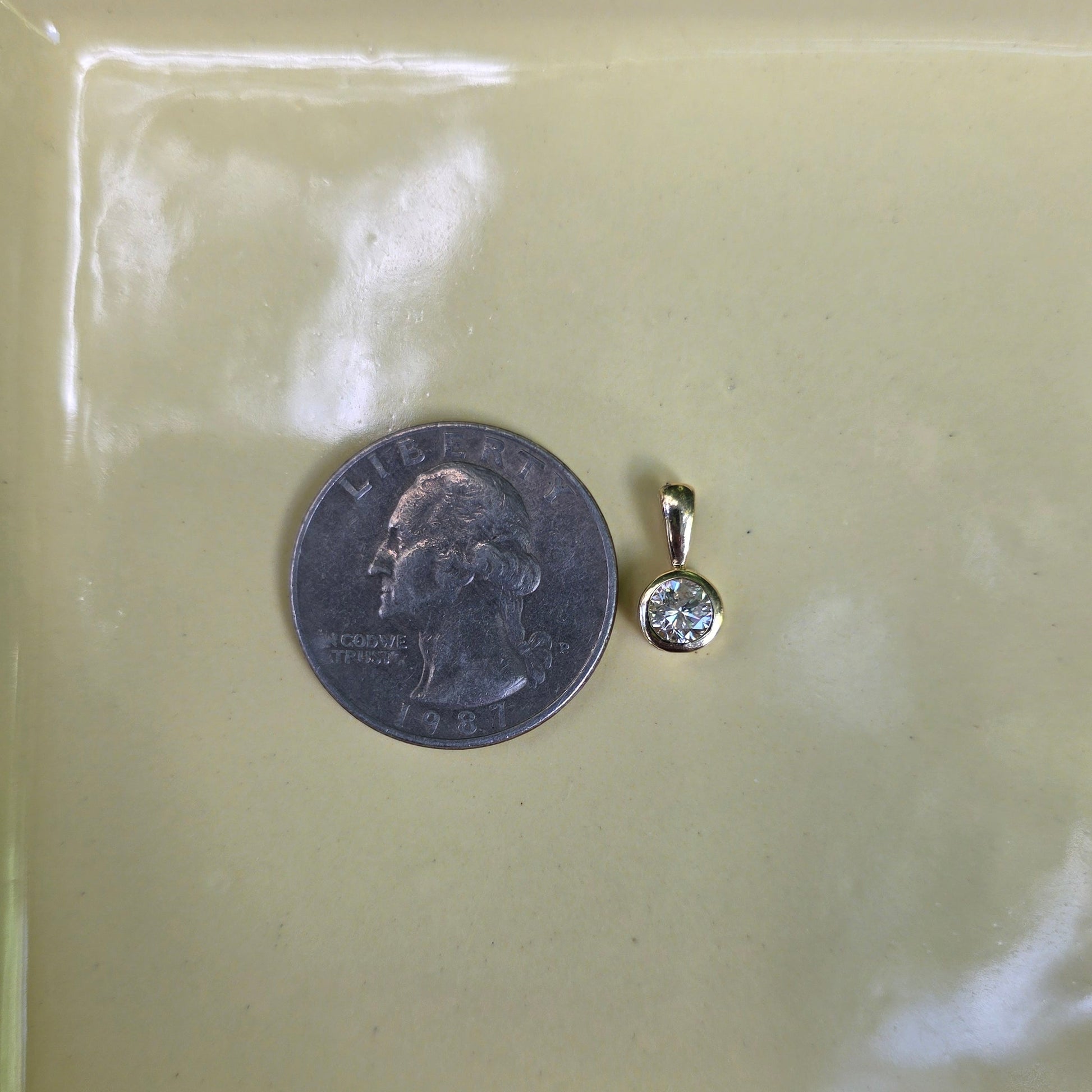 A small gold pendant with a clear stone is shown next to a US quarter for scale, placed on a pale yellow tray.