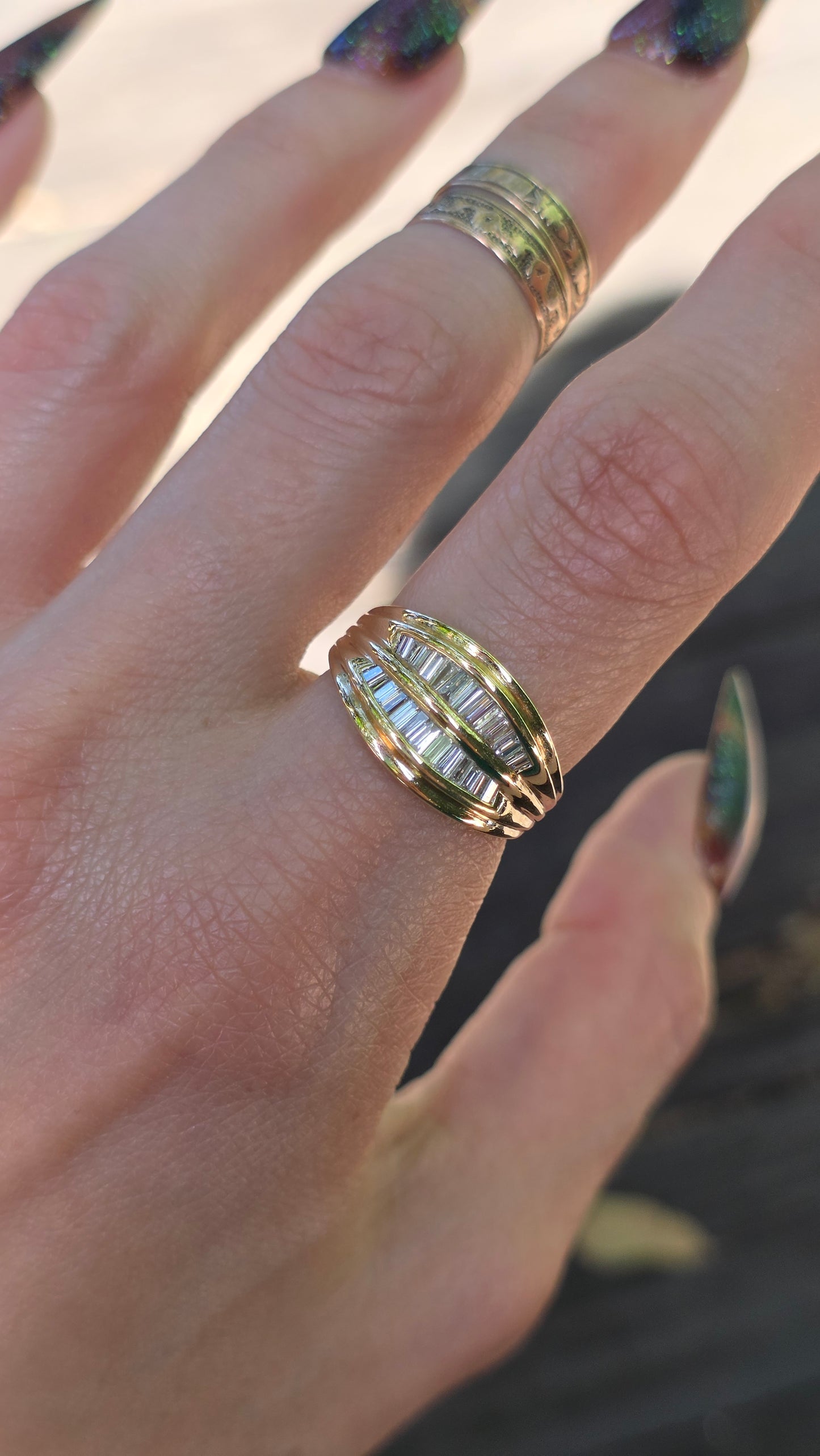 A close-up of a hand wearing a gold ring with baguette-cut clear stones, alongside other gold rings on other fingers.