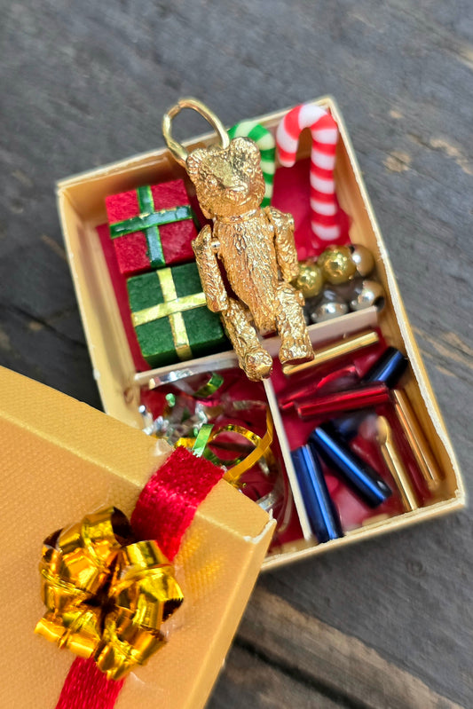 A gold teddy bear Christmas ornament displayed in an open gold gift box filled with miniature holiday items, with a gold gift box and red ribbon in the foreground on a dark wood surface.
