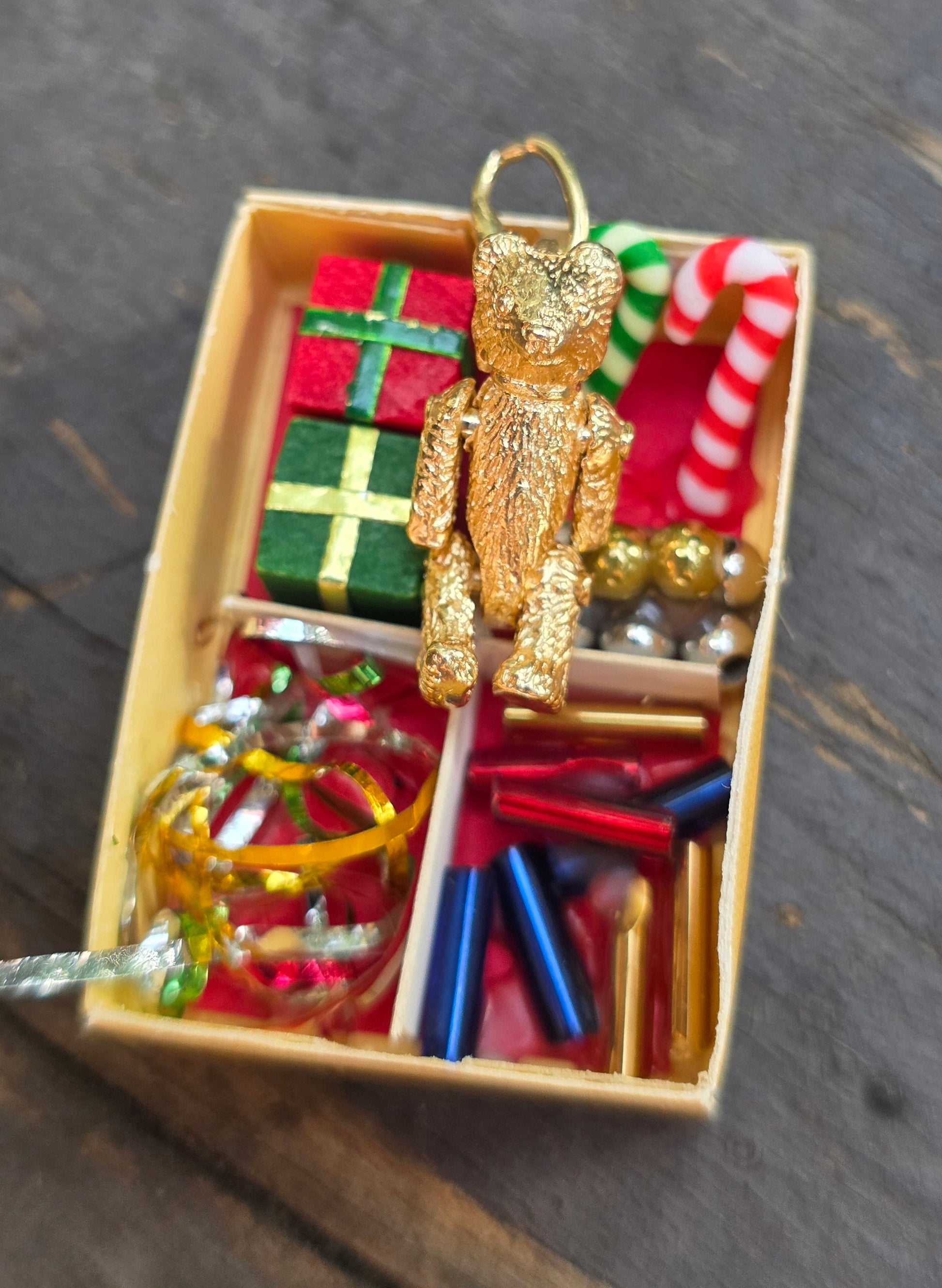 A small gold teddy bear ornament hanging above an open box of Christmas decorations, including wrapped gifts, candy canes, beads, tinsel, and metallic pieces, on a dark wooden surface.