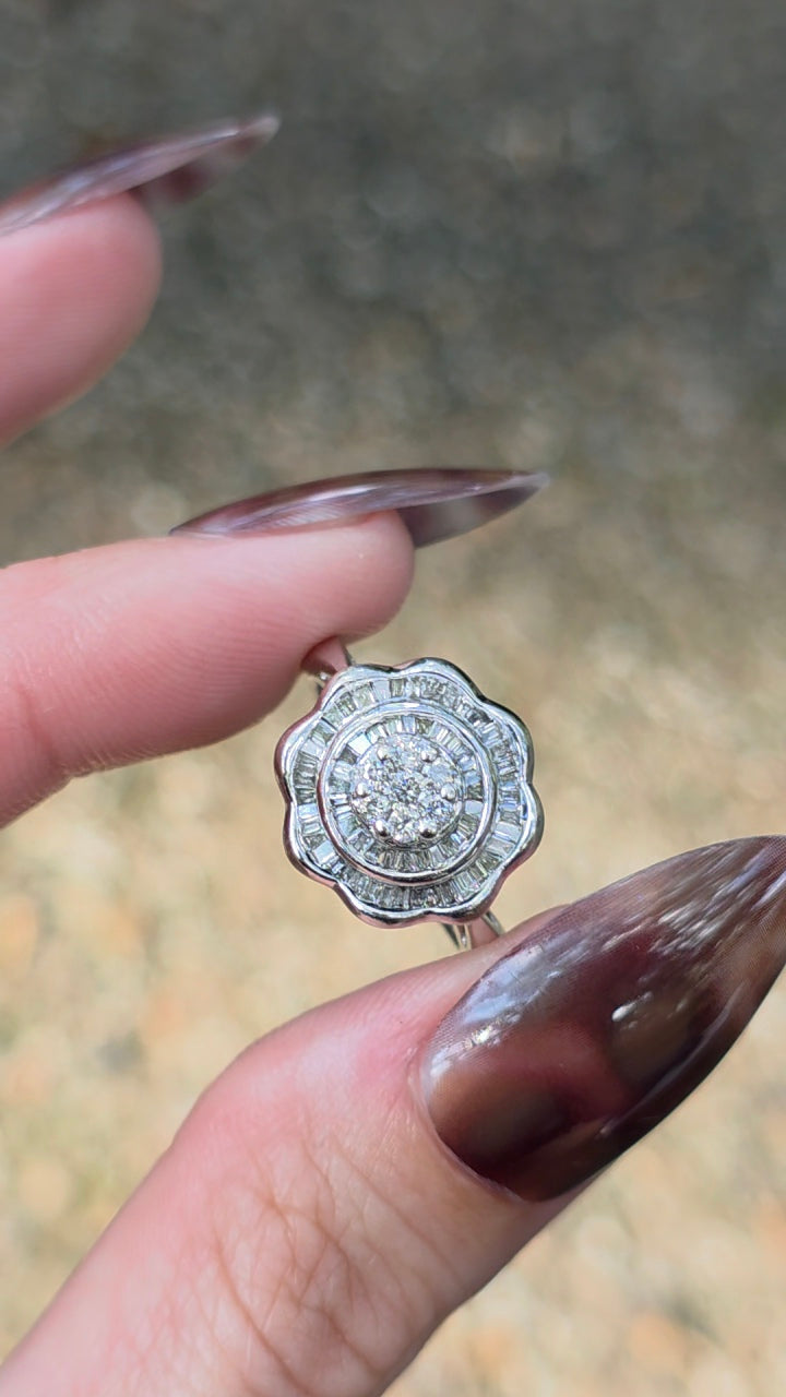 A close-up of a hand with long dark brown manicured nails holding a silver-toned ring featuring a round-cut clear gemstone surrounded by a halo of smaller clear stones.