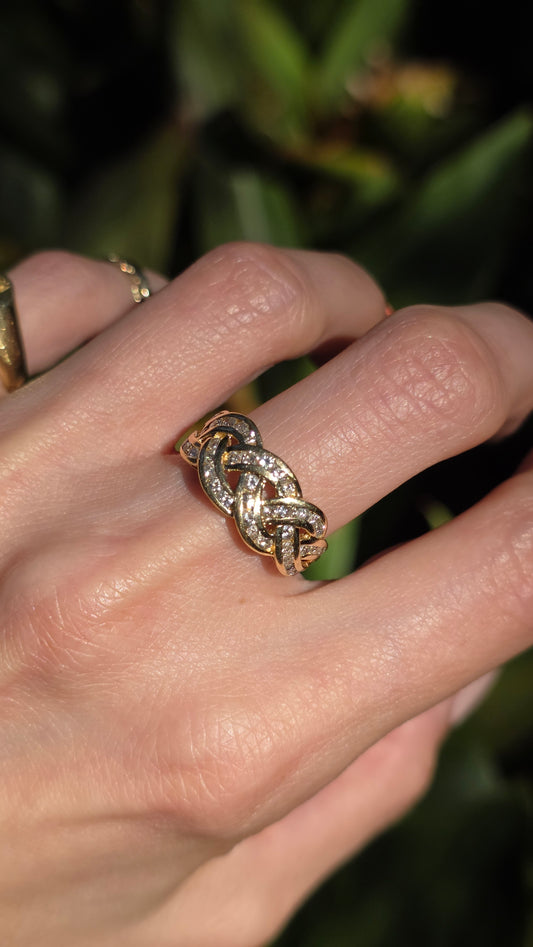 A close-up of a hand wearing a gold interlocking knot ring, with a blurred green outdoor background.