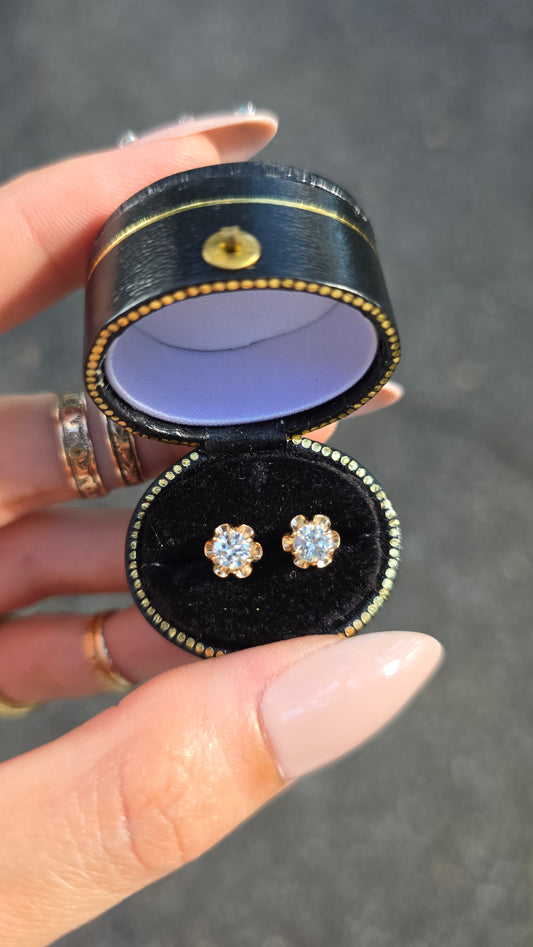 A pair of gold flower-shaped stud earrings with clear gemstones, displayed in an open black ring box held by a hand.