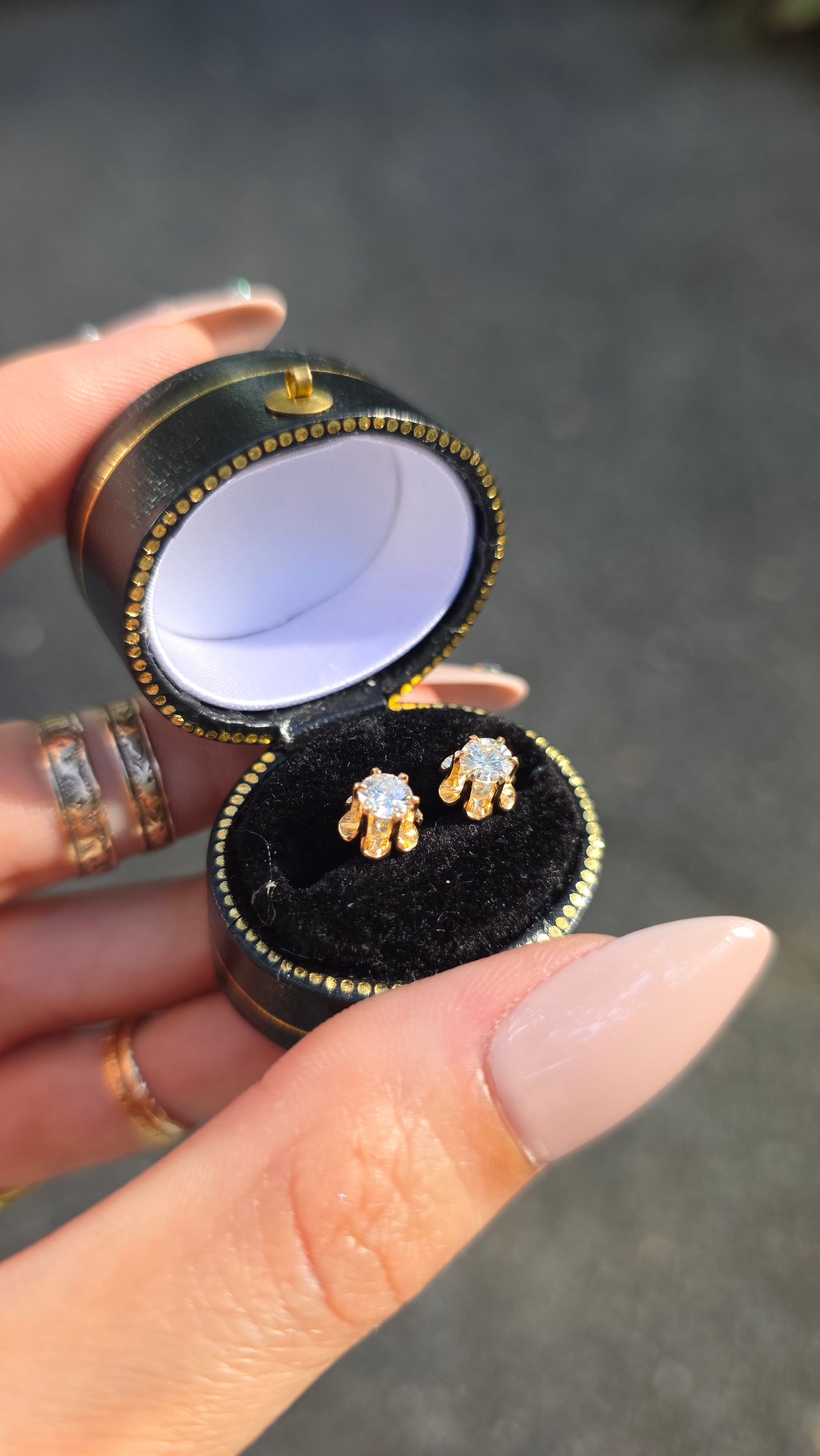 A close-up of a hand holding an open round black jewelry box with gold trim, showing a pair of gold earrings with clear stones set inside a black velvet insert.