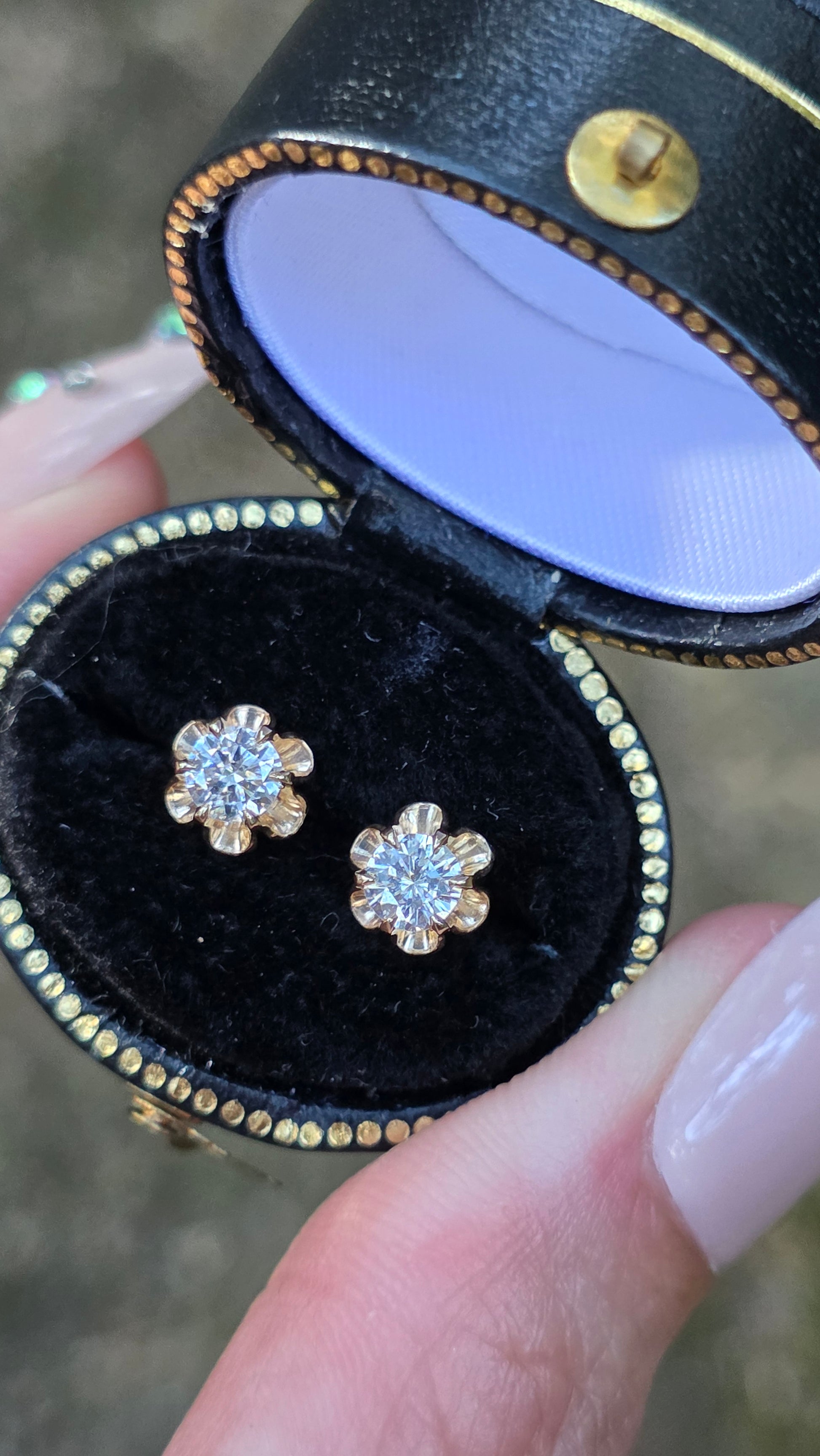 A close-up of a person's hand holding an open black jewelry box with a black velvet insert, displaying a pair of yellow gold flower-shaped stud earrings with clear gemstones.