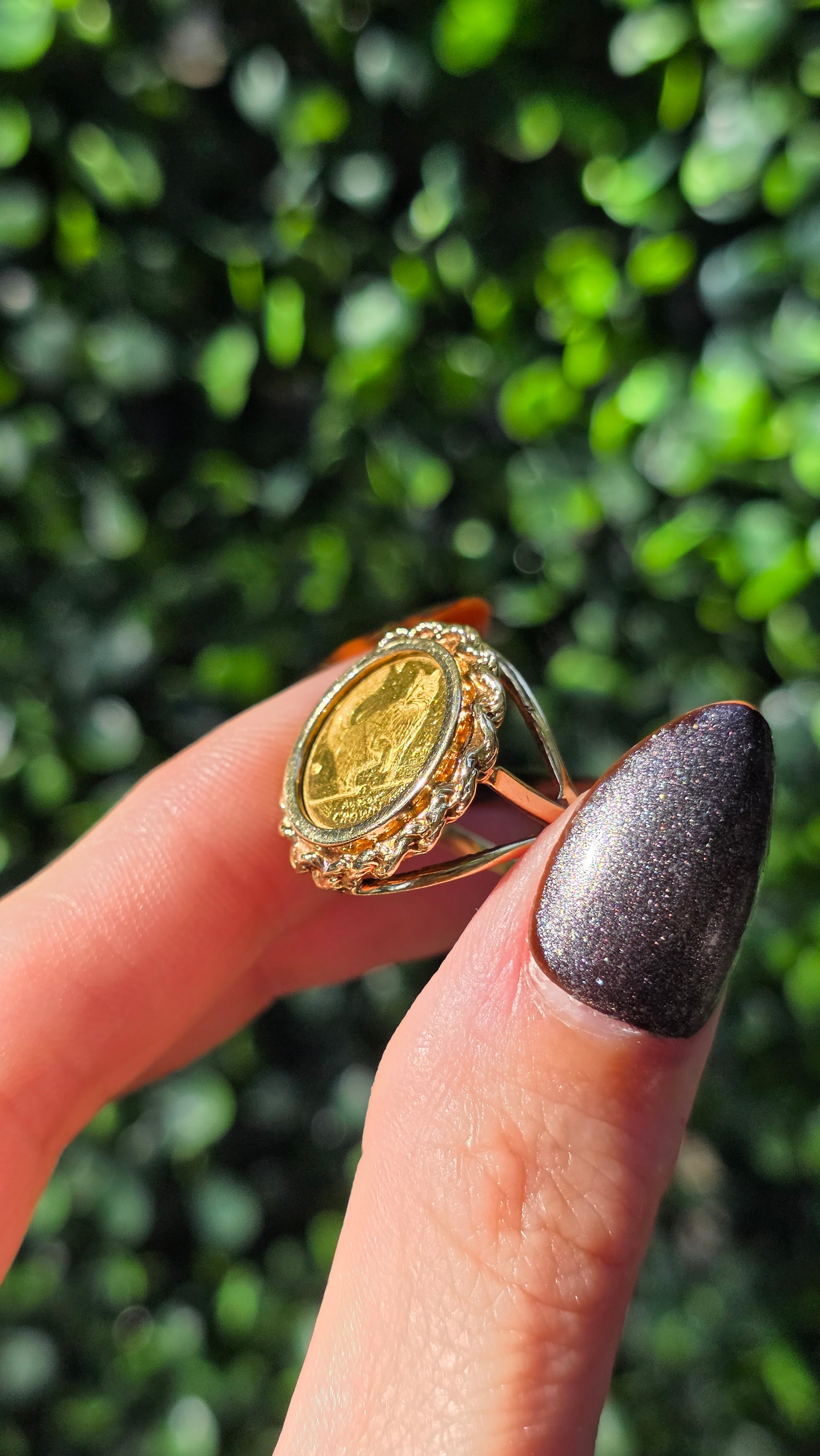 Gold ring with a coin design held by a finger against a blurred green background