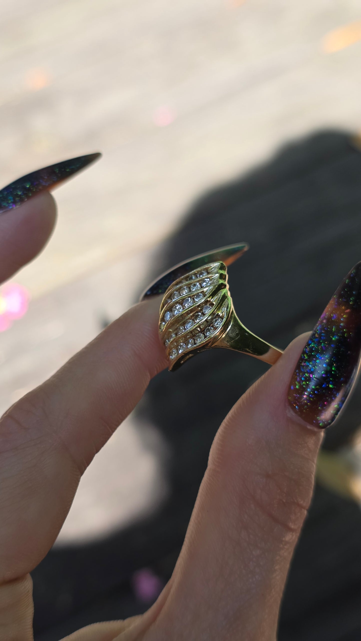 A gold ring with a curved, scalloped setting set with clear crystals, held between fingers with long, dark glittery manicured nails.