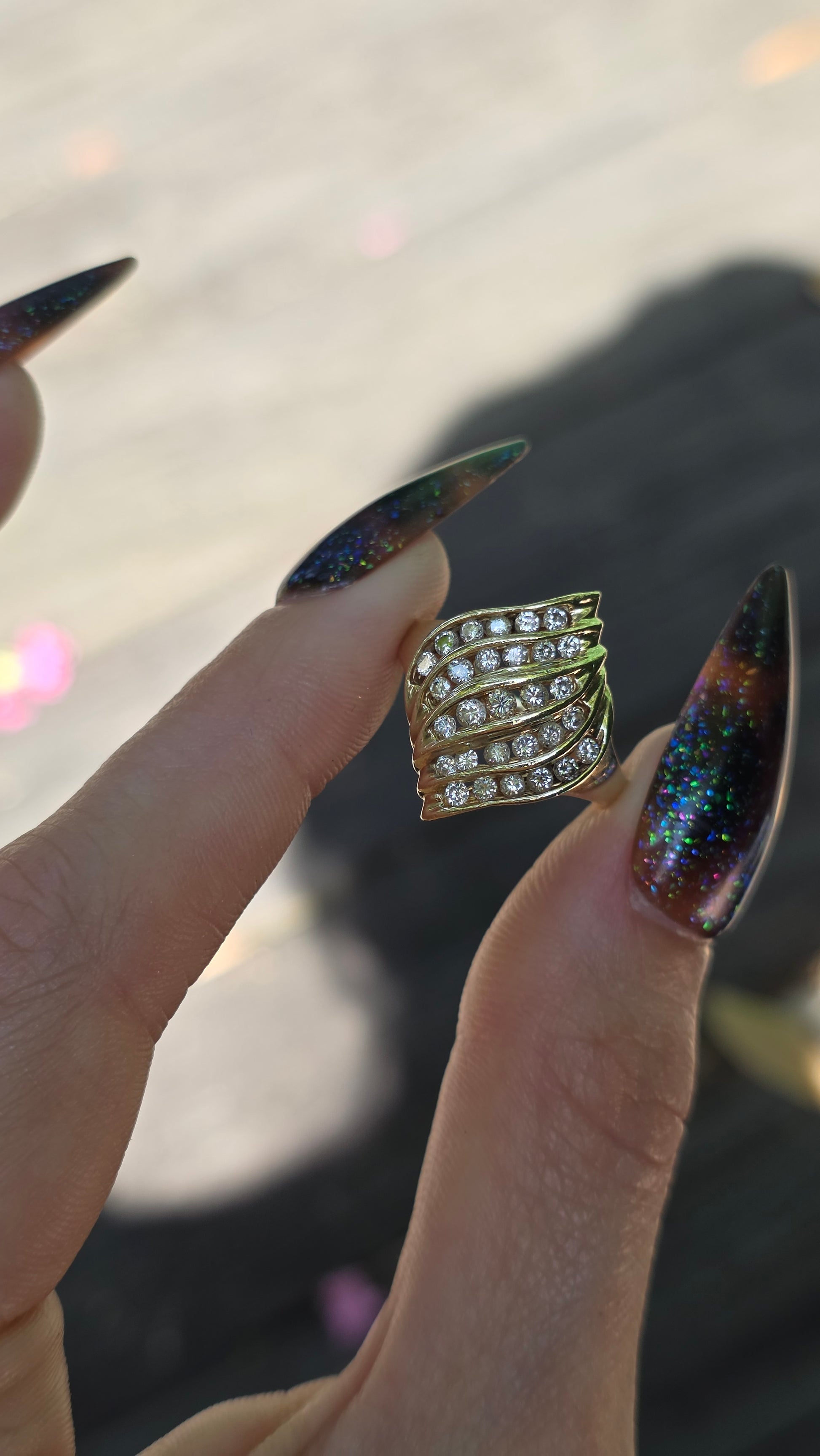 A gold ring with multiple rows of clear diamonds is held between two hands with long, dark glittery stiletto nails.