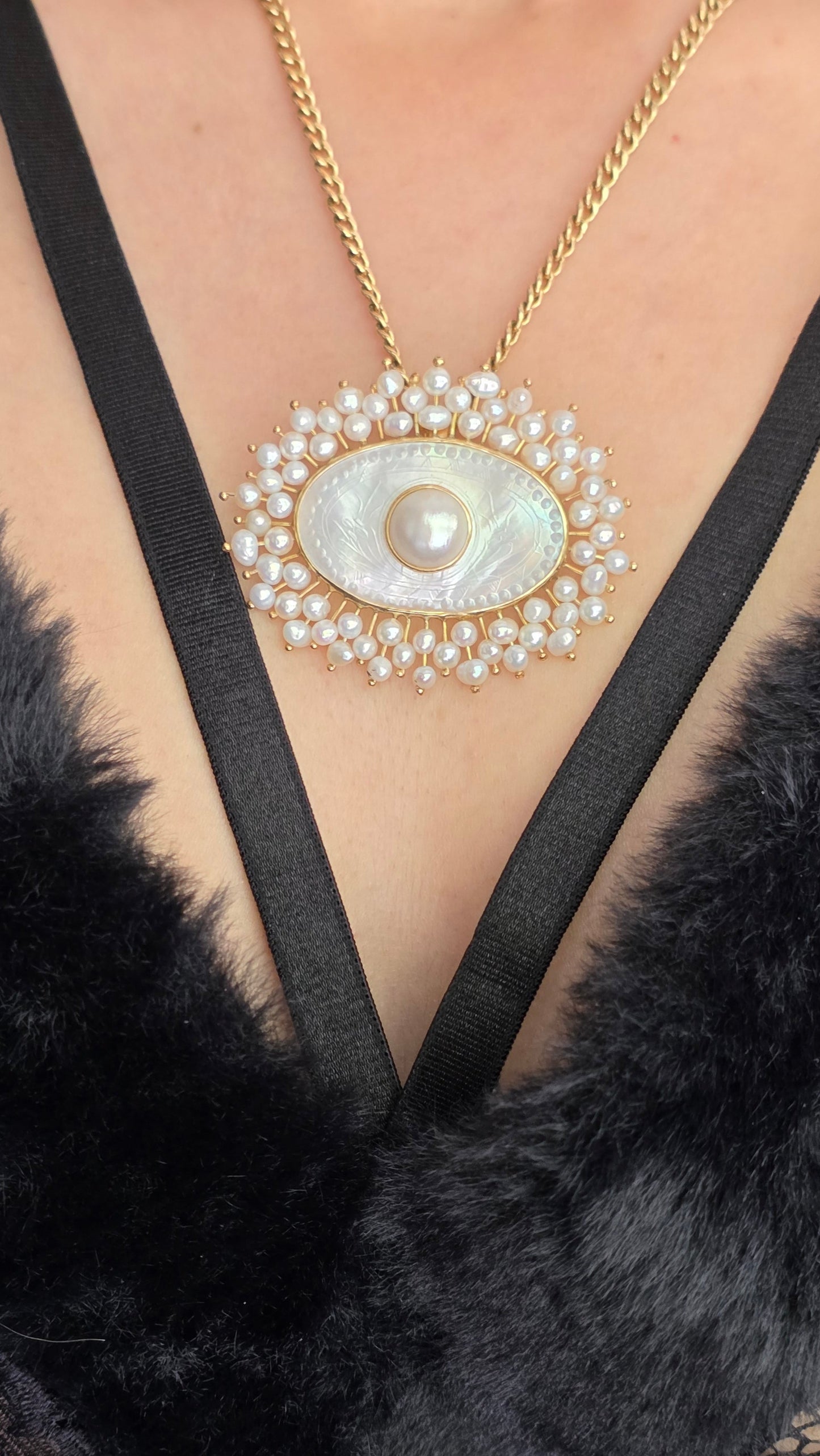 Gold necklace with an eye-shaped pendant on a person wearing a black fur garment.