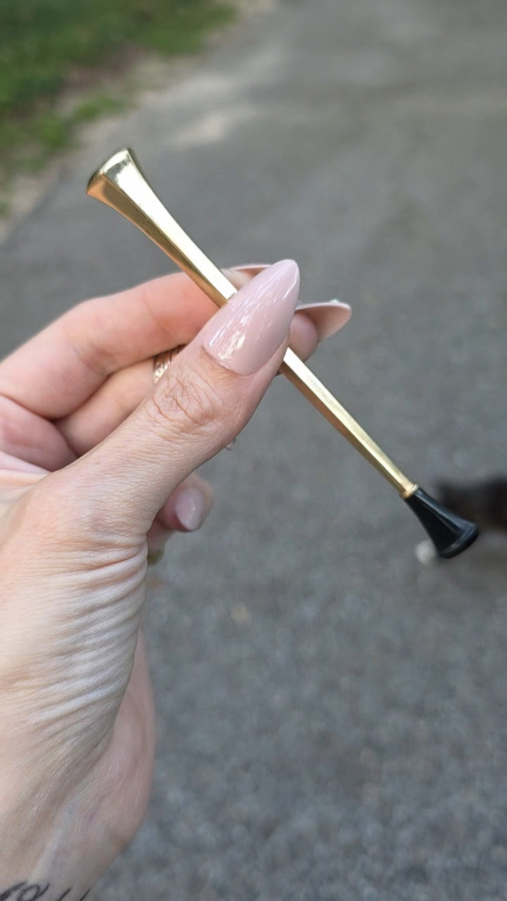 A hand with long nude manicured nails holds a slim gold metal tool with a black tip against an outdoor paved background.