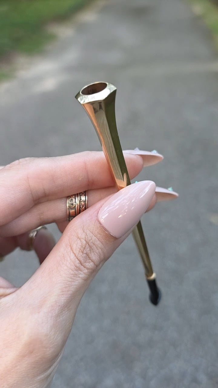 A hand with pale pink manicured nails holds a gold metal lip tool or makeup applicator with a black bristle tip against an outdoor blurred background of a paved path.