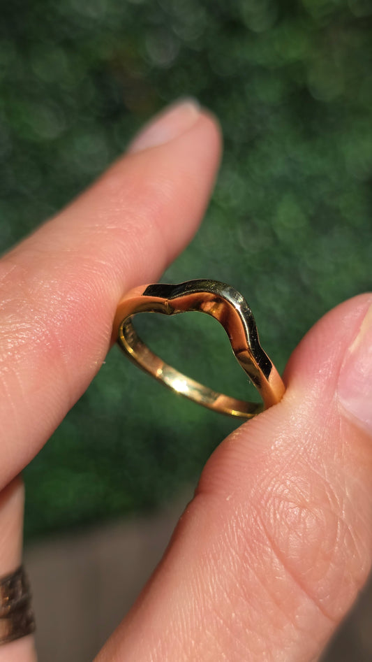 A close-up of a hand holding a gold ring with a wavy, organic silhouette against a blurred green background.