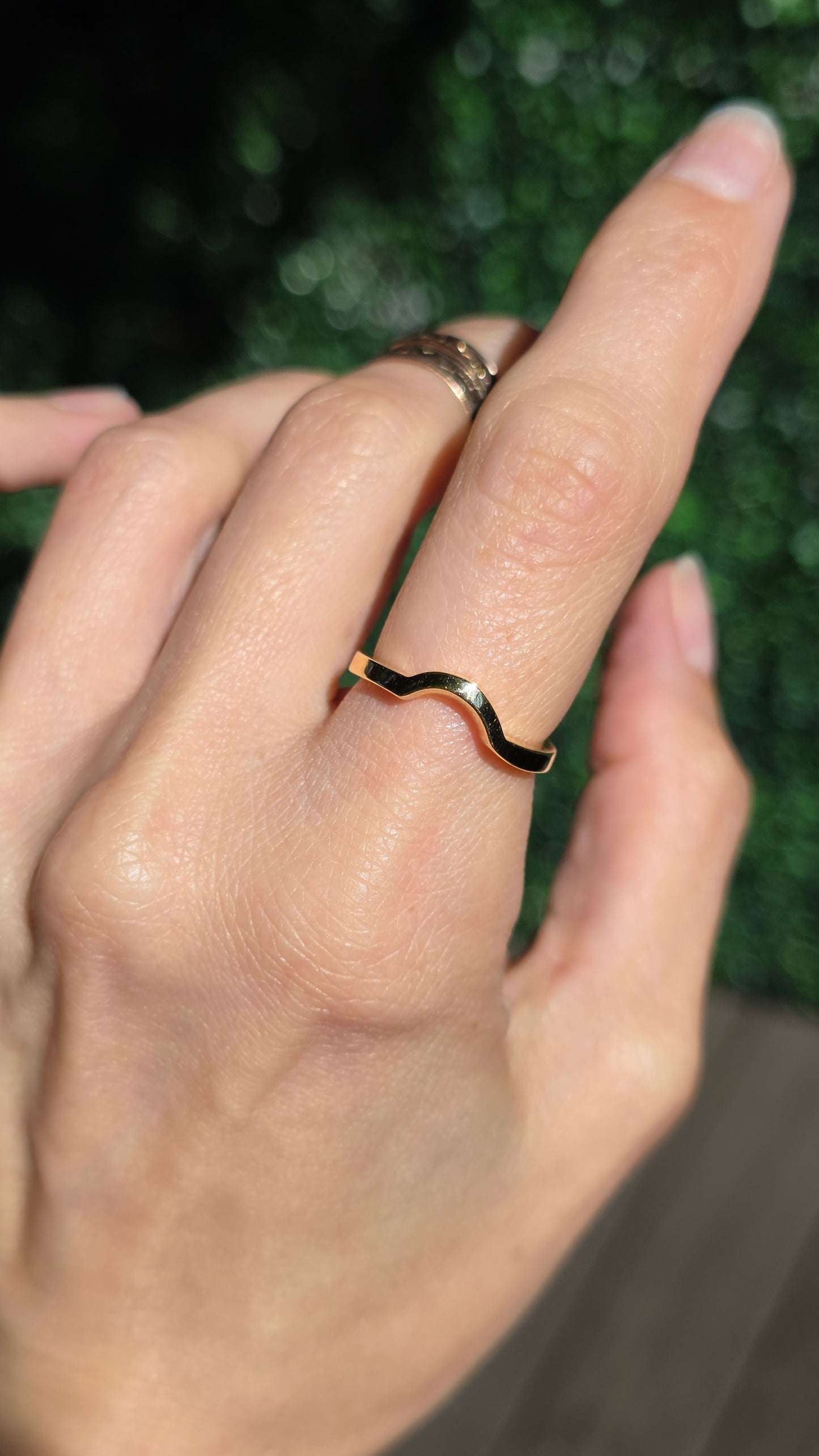 A close-up of a hand wearing a thin wavy gold ring on the ring finger, with a second silver-toned ring on the middle finger, against a blurred green background.