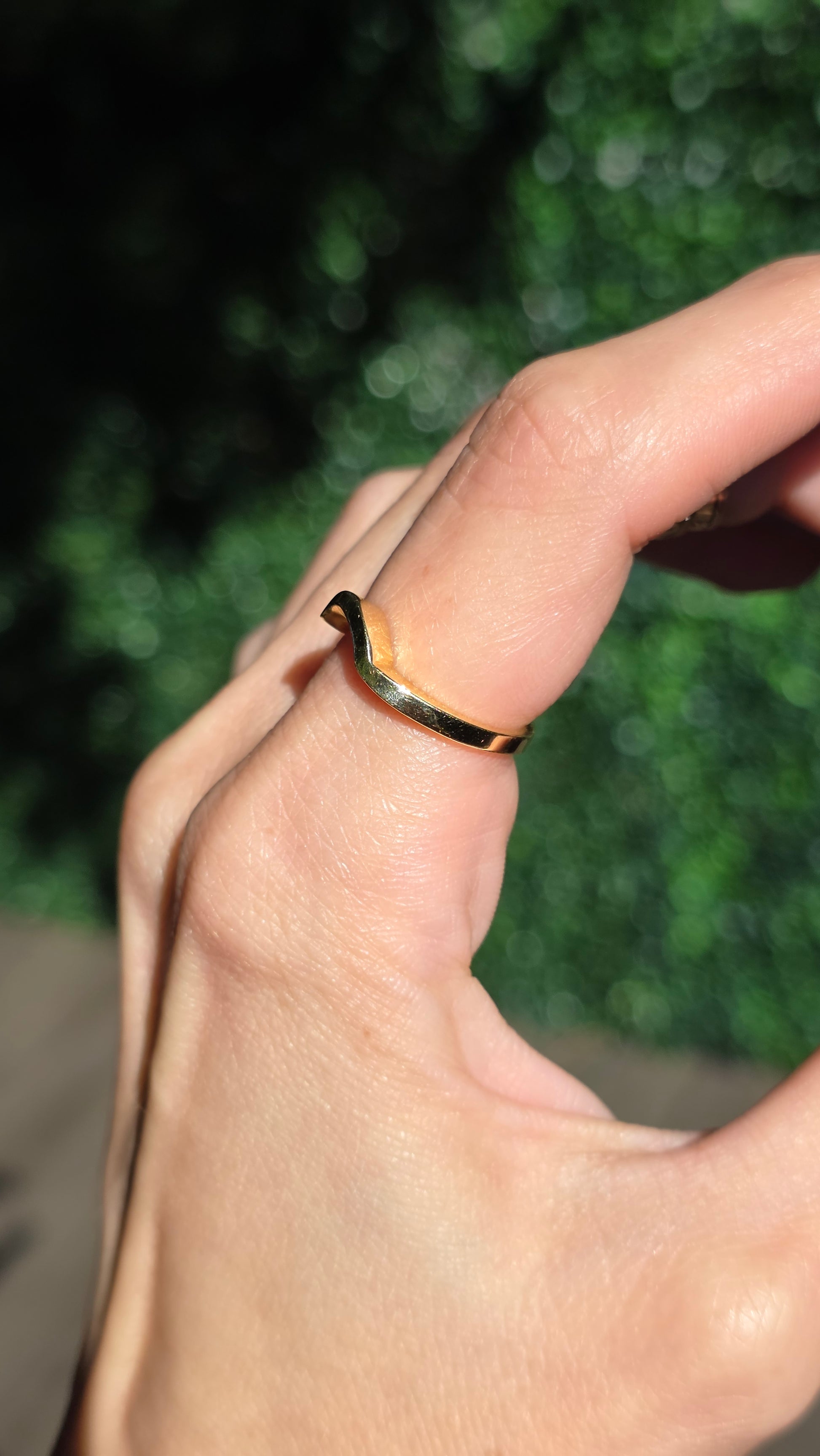 A close-up of a hand wearing a thin, twisted gold band ring against a blurred green outdoor background.