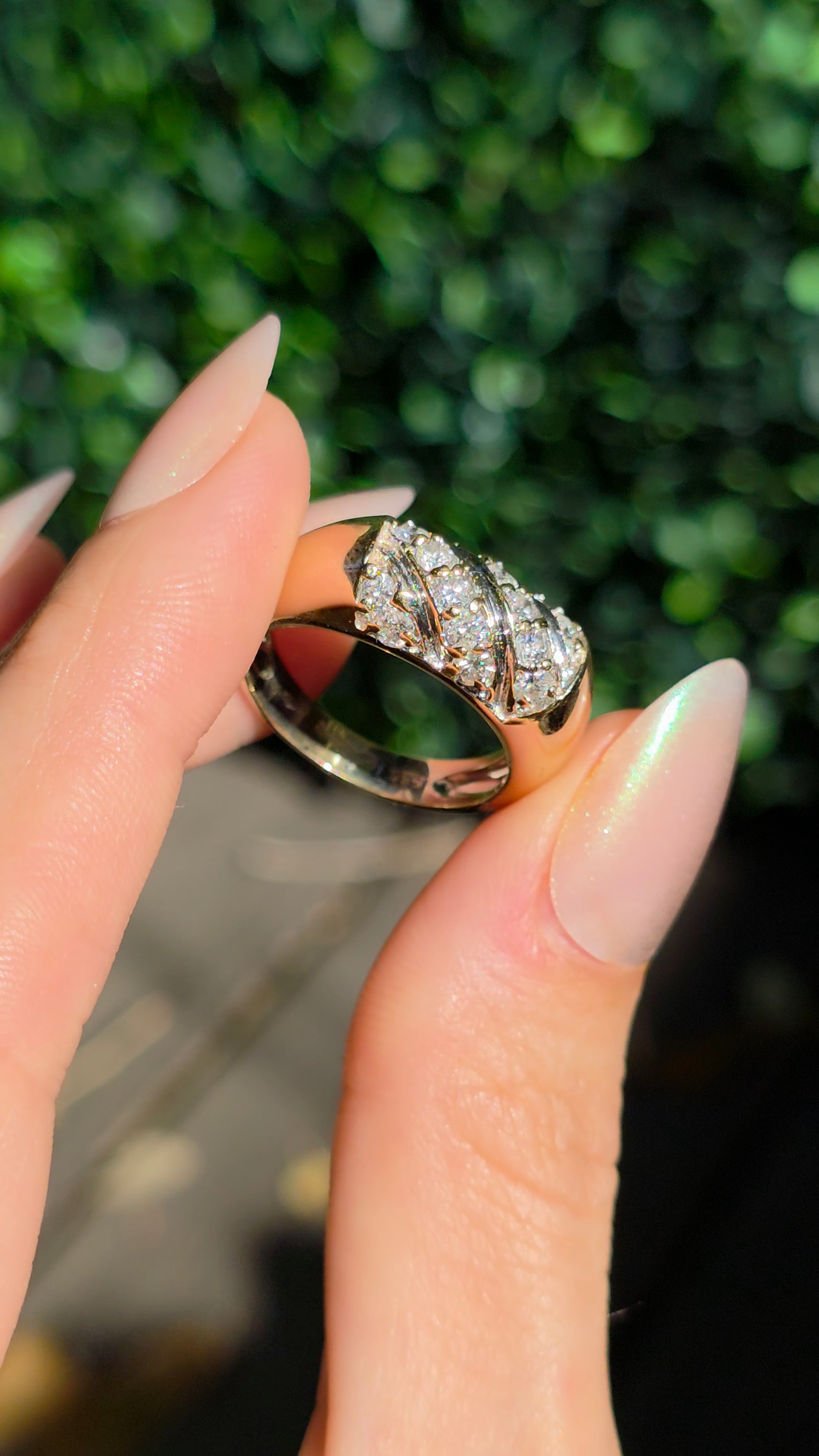 A close-up of a hand with manicured nails holding a silver ring featuring a cluster of clear gemstones on a rose gold band, with a blurred green background.