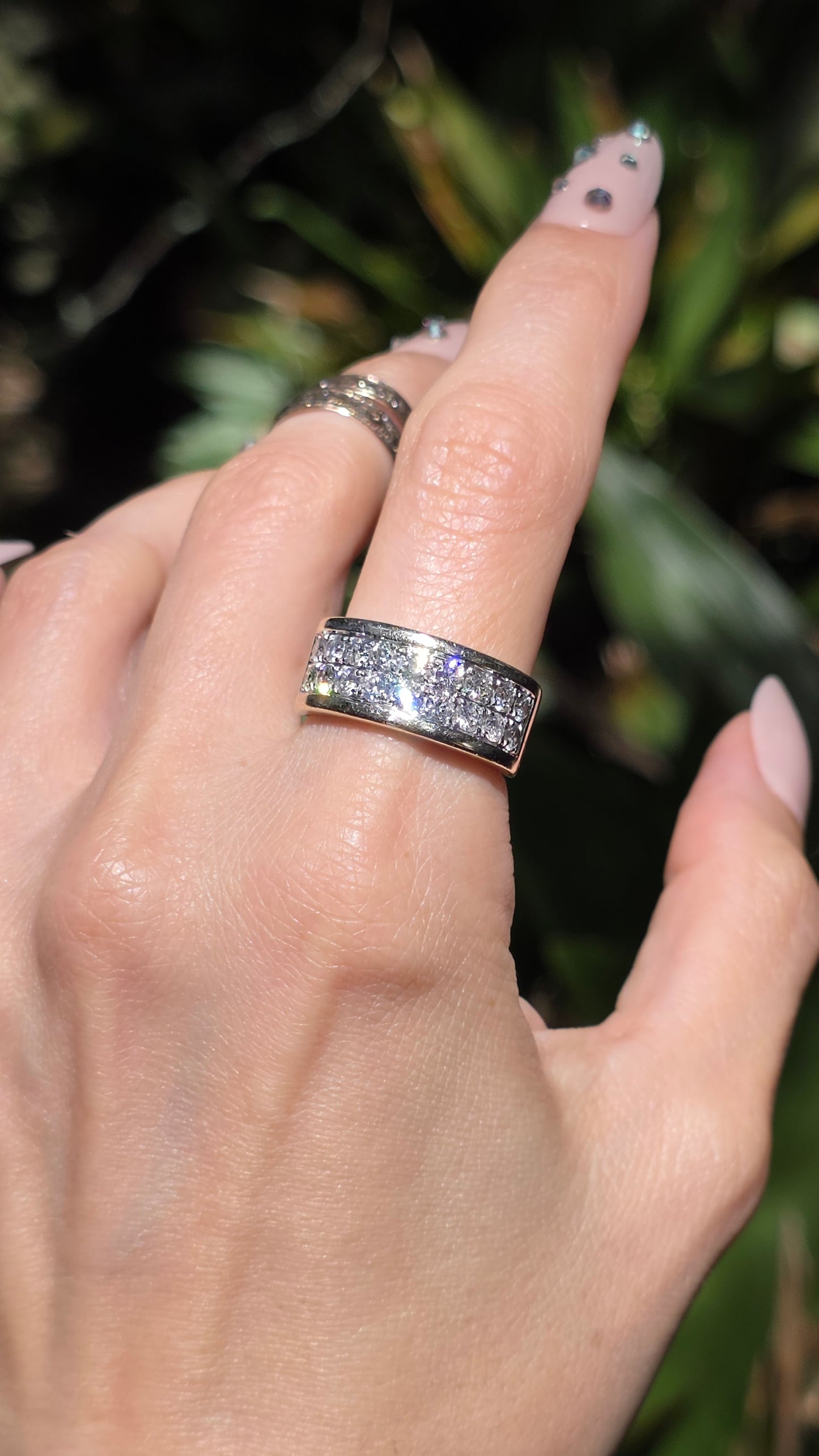 A close-up of a hand wearing a silver band ring set with clear pavé gemstones, with a second silver band ring on the middle finger, against a blurred green outdoor background.