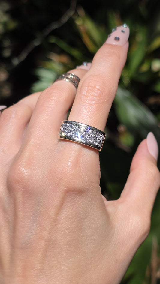A close-up of a hand wearing a silver band ring set with clear pavé gemstones, with a second silver band ring on the middle finger, against a blurred green outdoor background.