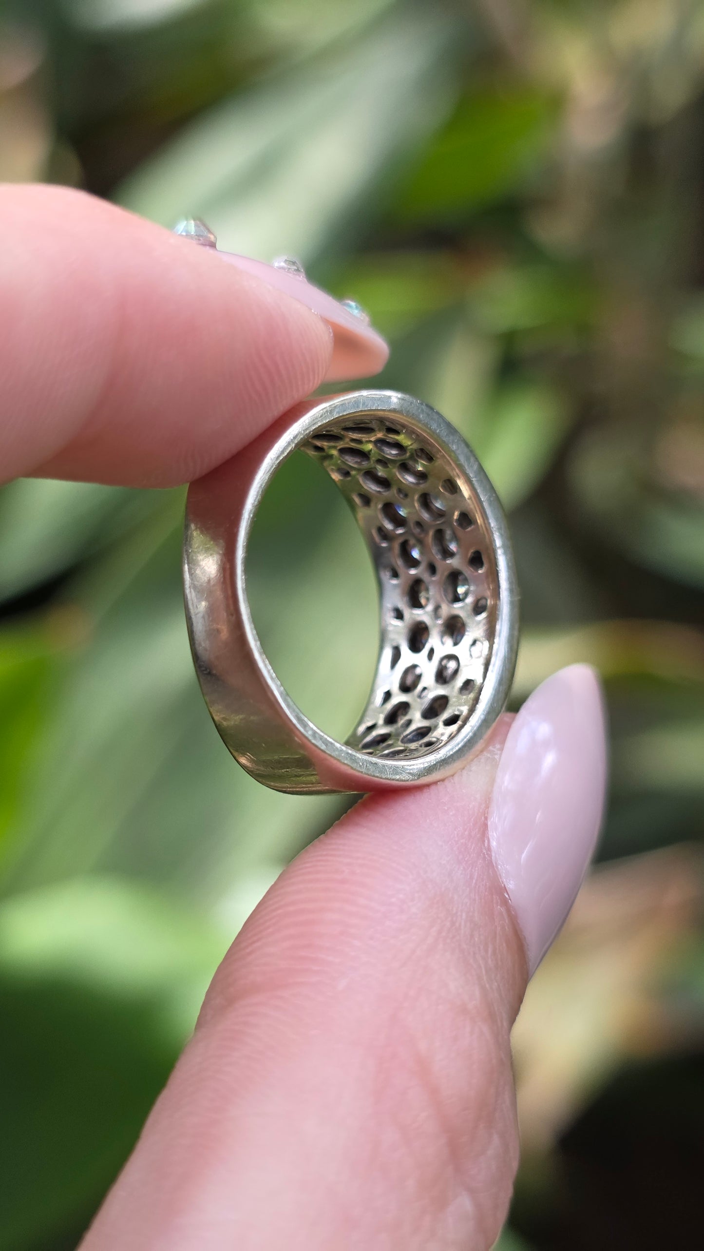 A close-up of a hand holding a silver ring with a textured, perforated inner band, set against a blurred green outdoor background.