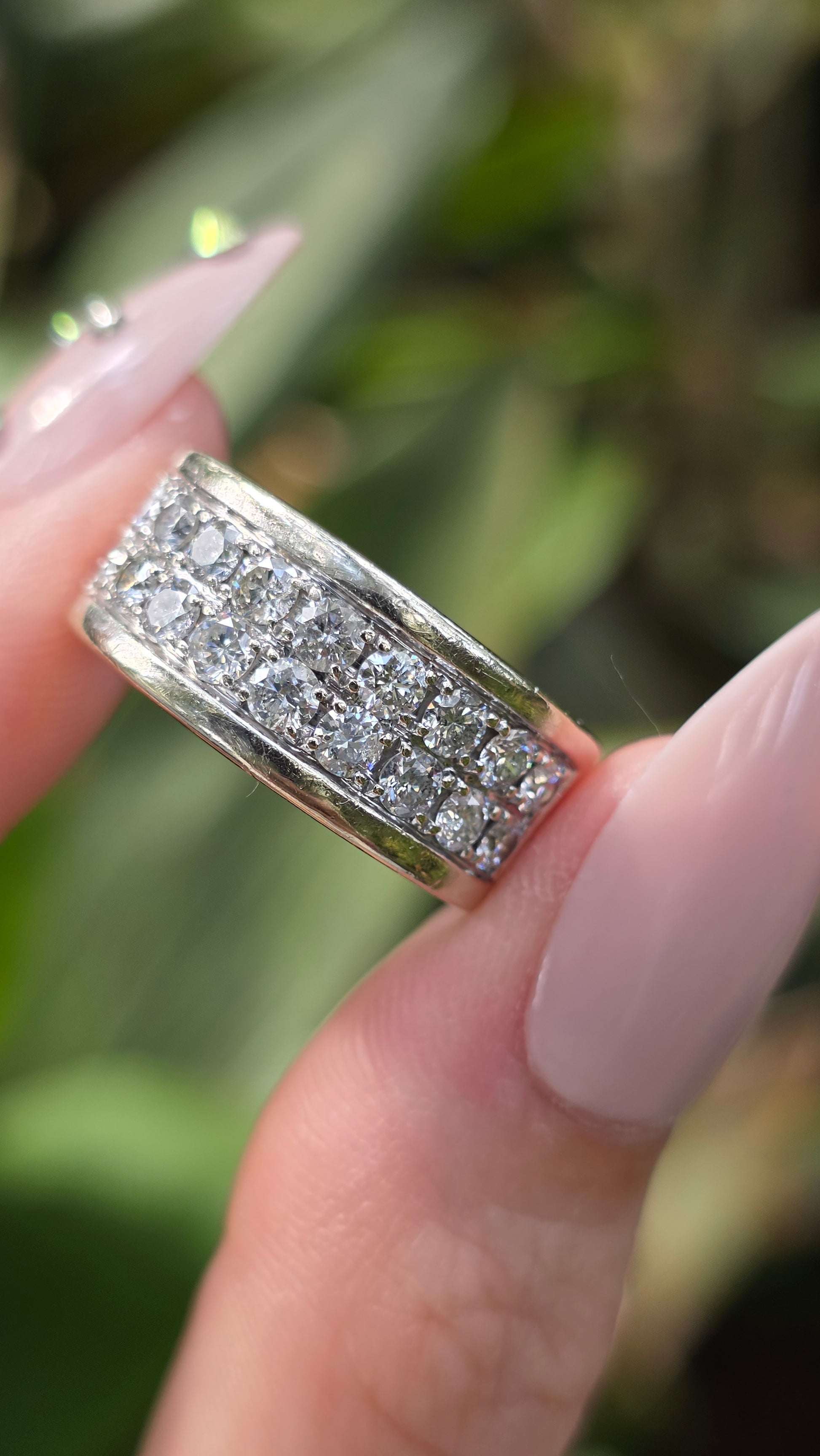 A close-up of a silver band ring with multiple rows of sparkling clear stones, held between fingers against a blurred green outdoor background.