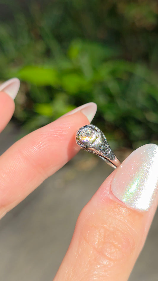 A silver ring with a teardrop-shaped clear gemstone held between two fingers against a blurred green outdoor background.