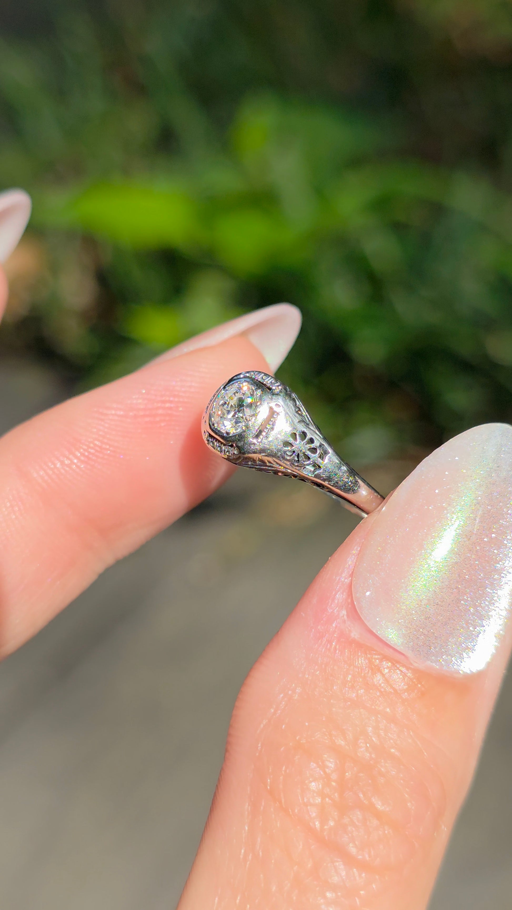 A close-up of a hand with iridescent nail polish holding a silver ring with a teardrop-shaped clear gemstone against a blurred green outdoor background.