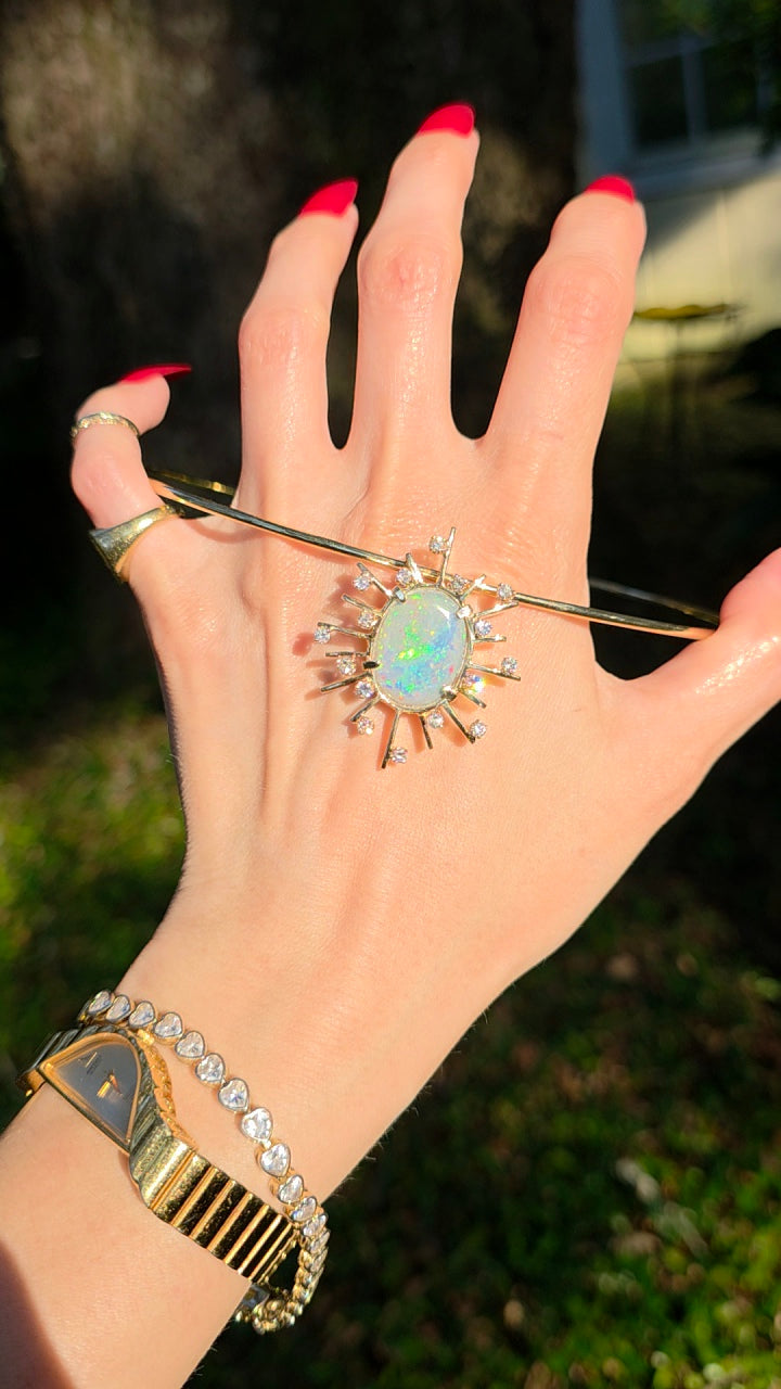 A gold-toned ring with a central oval opal surrounded by small clear stones, worn on a hand with red nail polish. A gold watch and bracelet are also visible on the wrist, with an outdoor garden background.