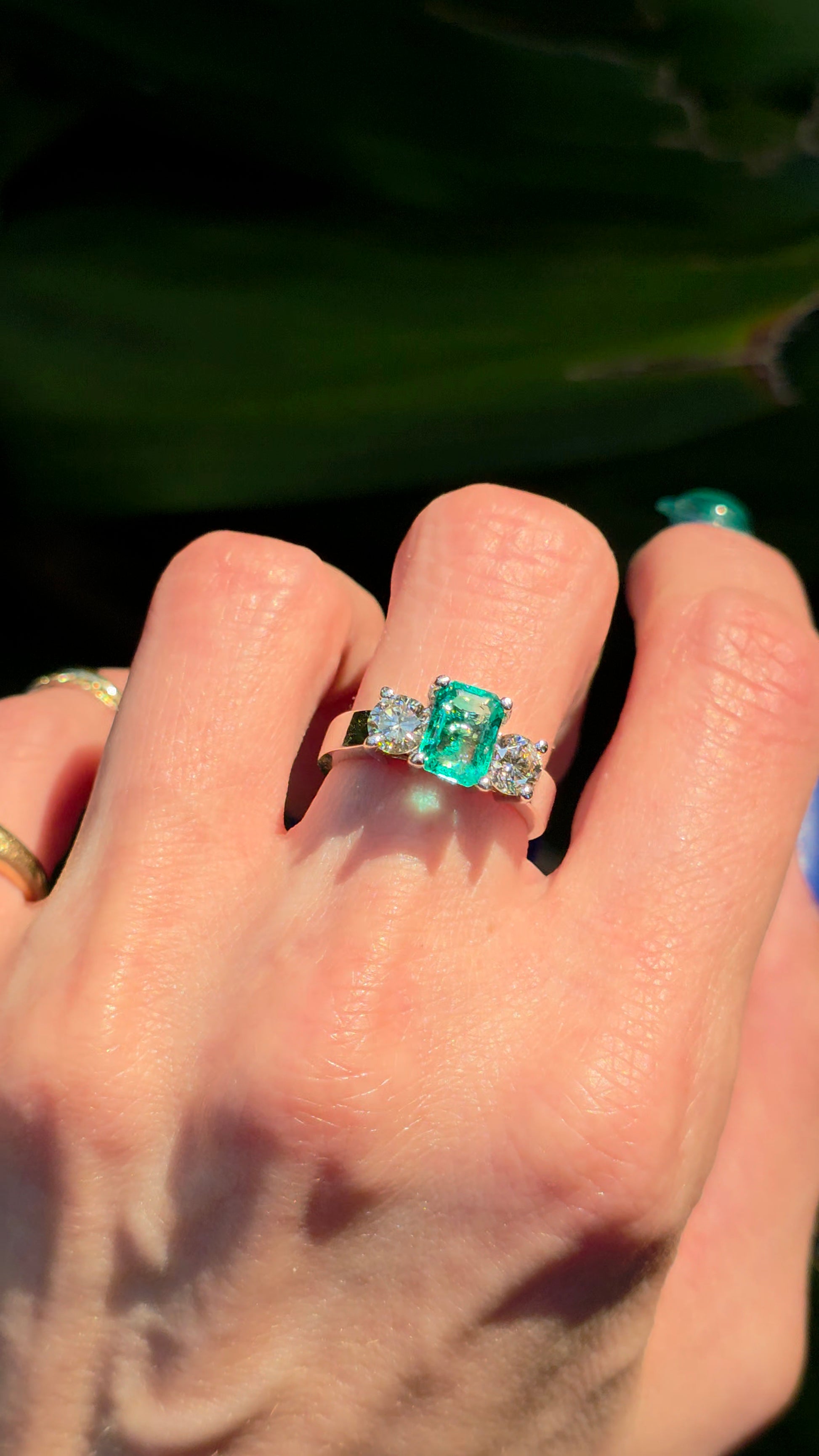A close-up of a hand wearing a silver ring with a rectangular green emerald-cut center stone and small clear side stones, photographed outdoors with a dark green leafy background.