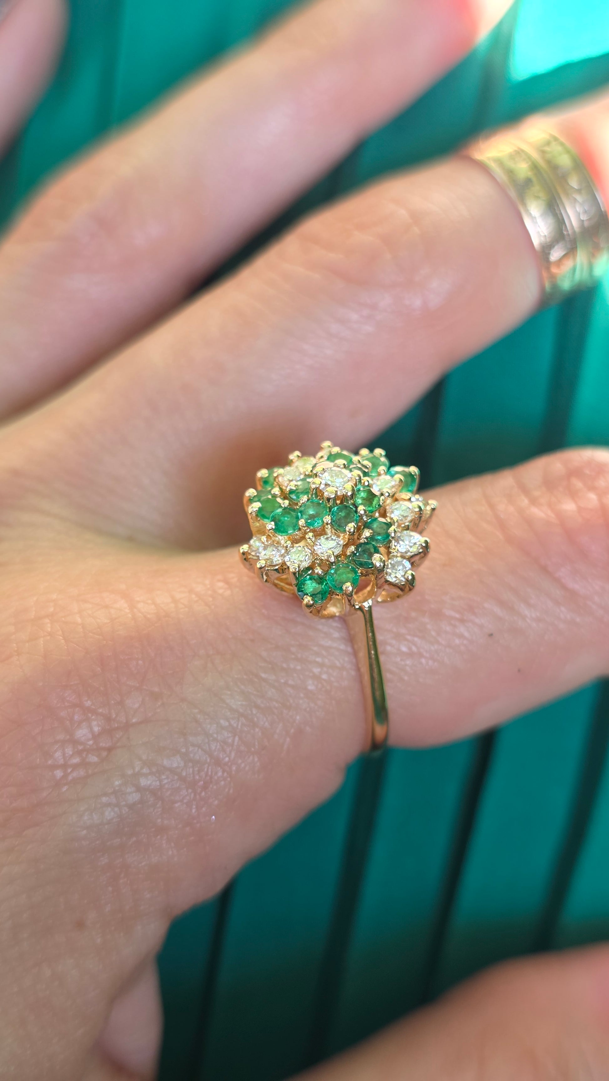 A close-up of a hand wearing a gold ring with a cluster of green and clear stones, with a second gold ring on the adjacent finger against a green background.
