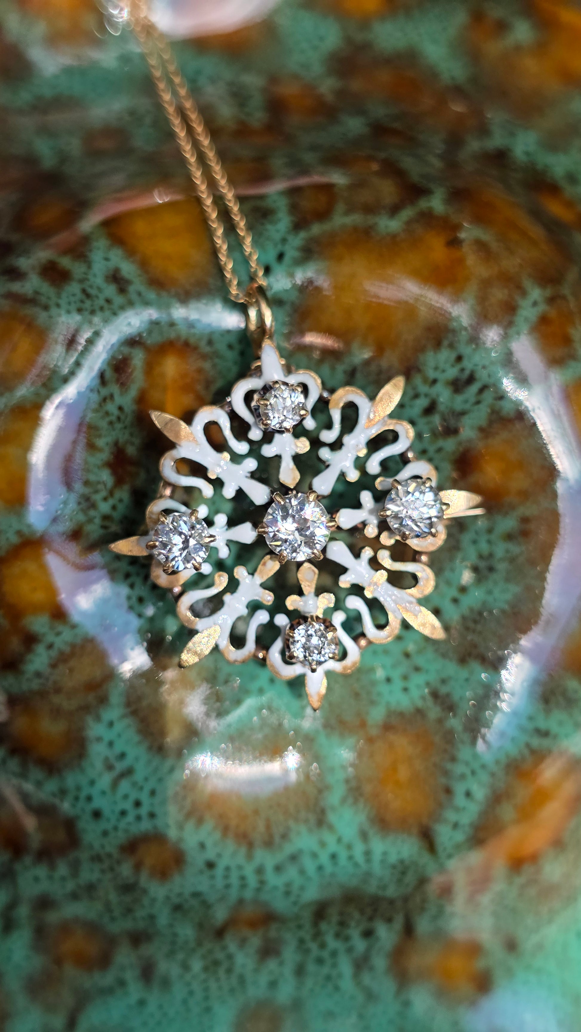 A close-up of a snowflake-shaped pendant necklace with clear gemstones and gold-toned filigree details, hanging against a glossy green and brown marbled surface.