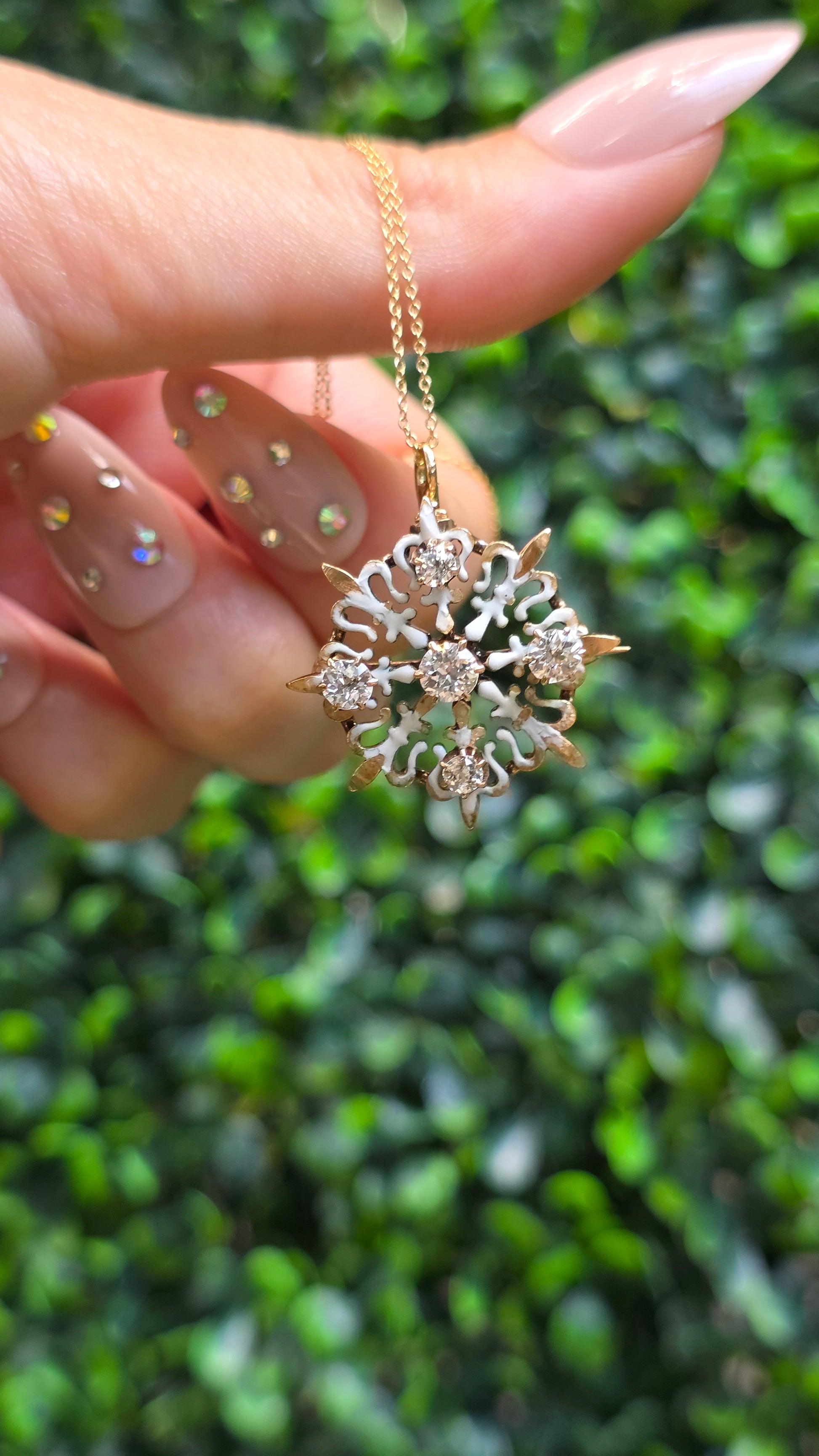 A gold snowflake pendant necklace with clear crystals, held by a hand with manicured nails against a blurred green outdoor background.