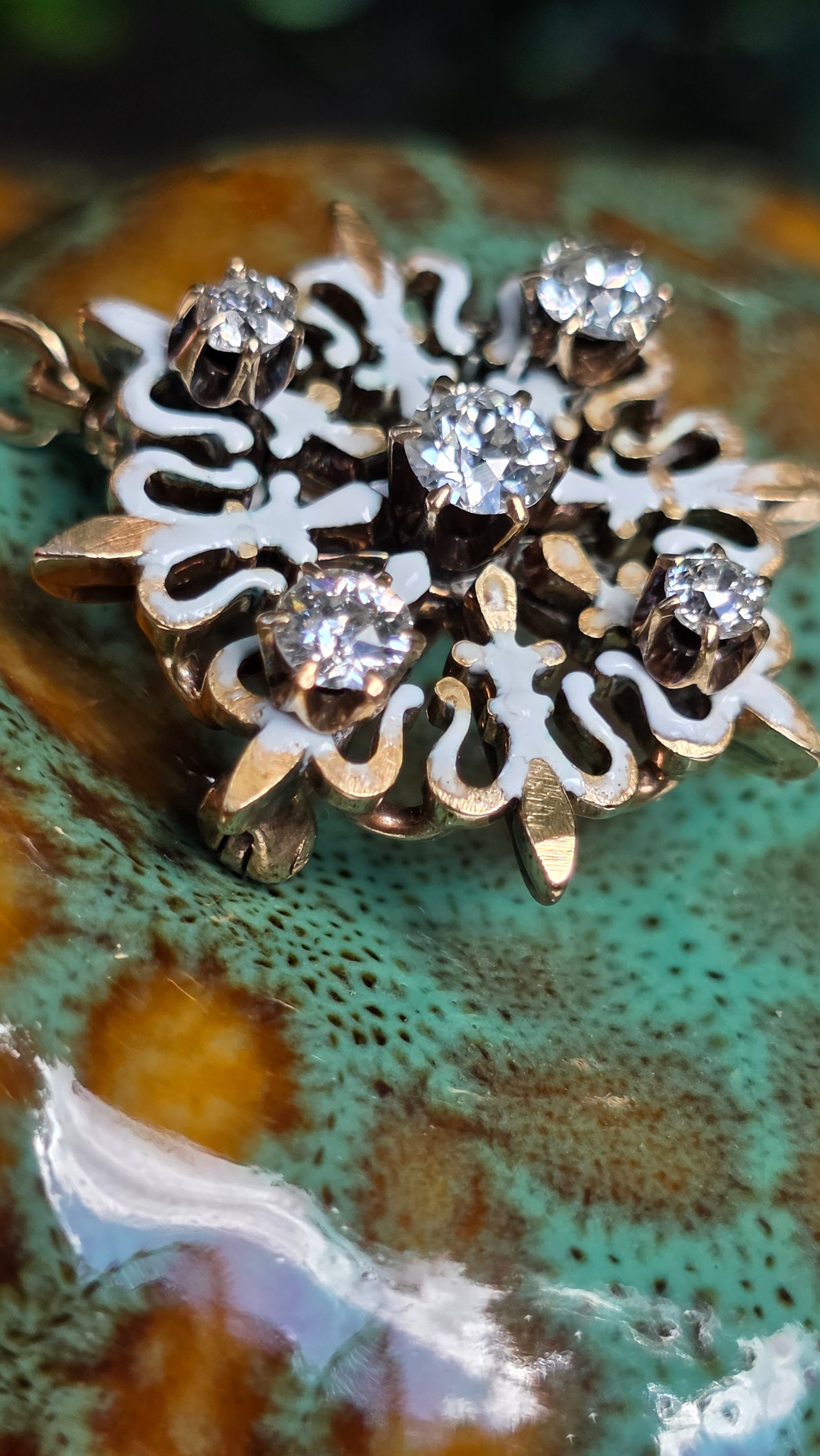 A close-up of an ornate silver-toned pendant with five clear round-cut gemstones and intricate filigree details, resting on a textured green and brown ceramic surface.