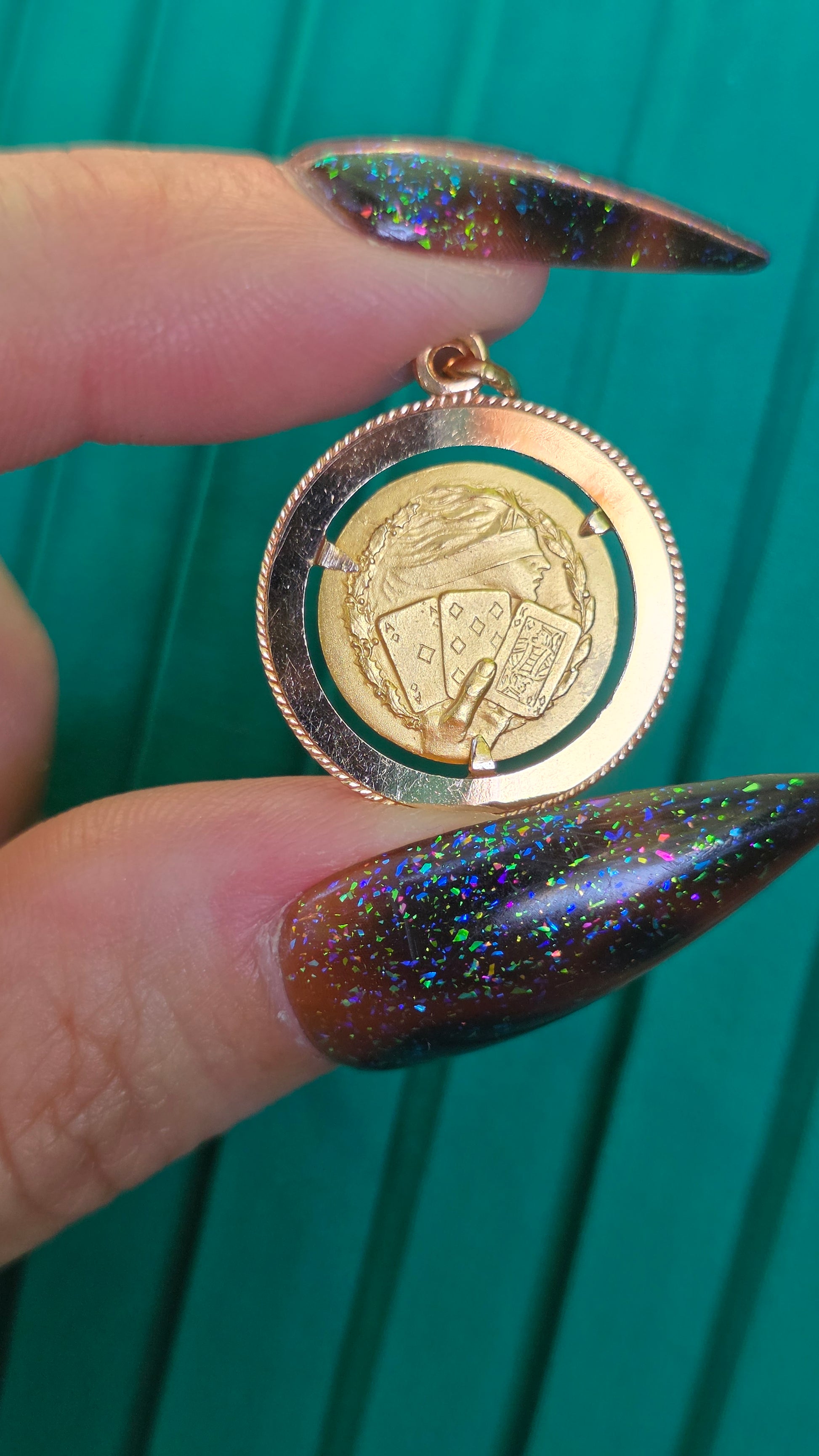 A close-up of a person's fingers with glittery dark nails holding a round gold pendant or locket with a green inner background and an embossed illustration.