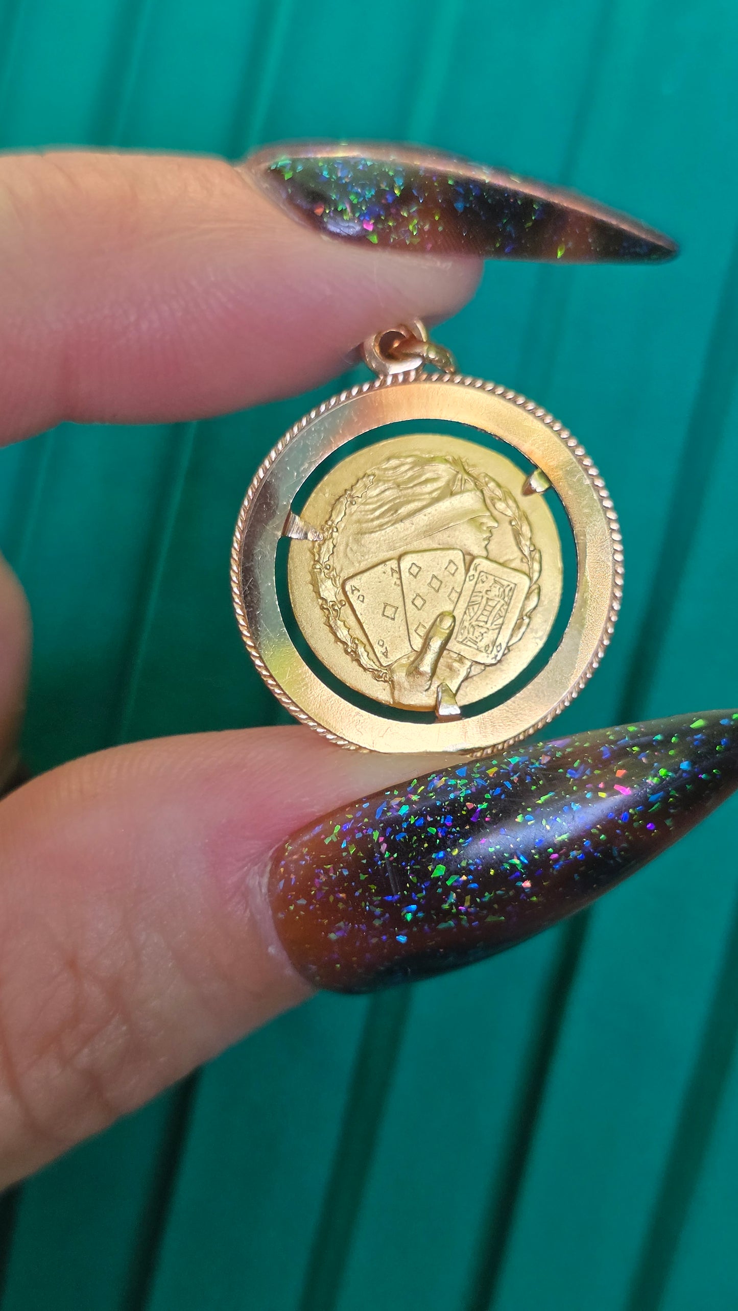 A gold-toned oval pendant or locket with an embossed crest design, held between two long glittery dark brown nails against a teal green background.