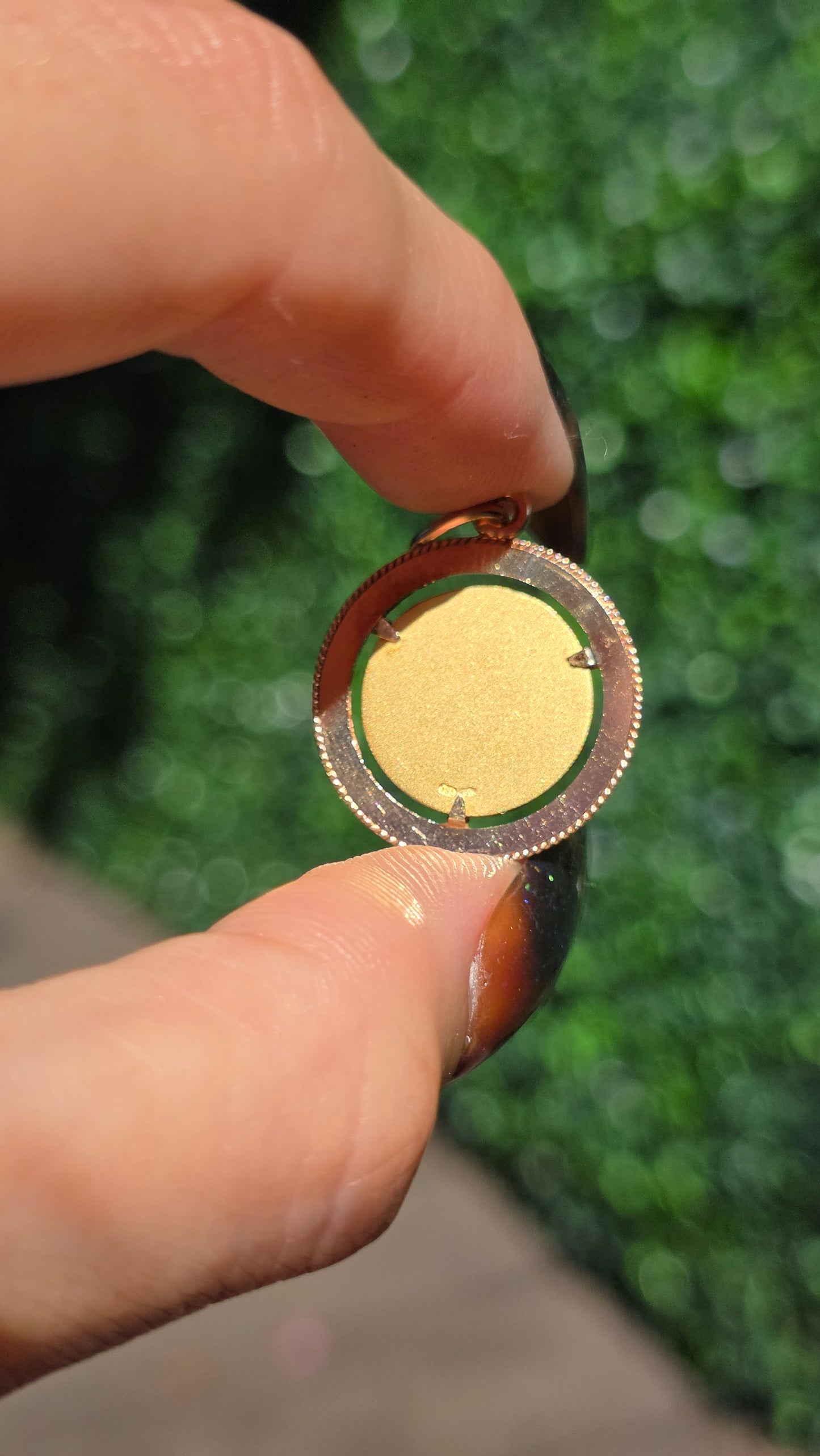 A close-up of a person's fingers holding a round gold-toned ring with a textured, beaded outer edge against a blurred green outdoor background.