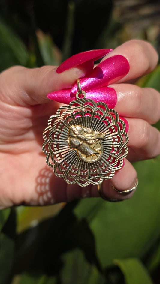 Gold pendant held by a hand with pink nail polish against a blurred green background
