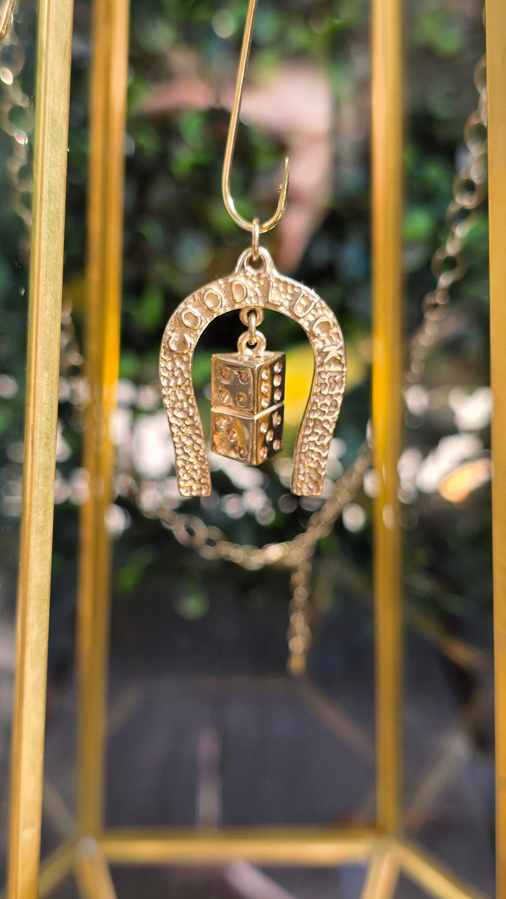 A gold-toned horseshoe-shaped pendant with a small rectangular charm featuring a raised '1', hanging in front of a blurred gold display case and green foliage background.